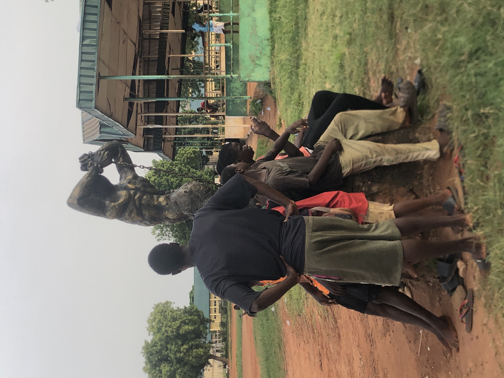 This image, taken in Nsugbe, Nigeria, captures an outdoor scene featuring several individuals gathered around a prominent statue on a mixed dirt and grassy field.
**Subjects:** The primary subjects are a group of dark-skinned individuals, mostly young males, dressed in casual attire such as t-shirts, shorts, and trousers. Some are lying or sitting on the reddish-brown earth and green grass, appearing relaxed. One person in a dark blue t-shirt and olive shorts stands with his back to the camera, hands on his hips. Another person is seen with hands raised, looking upward. A notable object is a large, dark-colored statue, possibly bronze, located to the left of the group. It depicts a powerful male figure with chains on his wrists and neck, seemingly in the process of breaking free. In the background, there are various buildings with light-colored walls and green or blue corrugated metal roofs, along with scattered green trees.
**Activity or event taking place:** The individuals appear to be resting, observing, or perhaps socializing in a relaxed manner. Their postures suggest a period of downtime or casual gathering. The presence of the statue might indicate a place of historical or cultural significance.
**Notable details:** The scene unfolds during the daytime under what appears to be an overcast or hazy sky, providing soft, diffused lighting rather than direct sunlight. The ground consists of both dry, reddish-brown dirt and patches of green grass. The statue's theme of breaking chains suggests symbolism related to freedom, struggle, or historical events. The architectural style of the background buildings and the reddish soil are consistent with the location context of Nigeria. There is no visible text in the image.