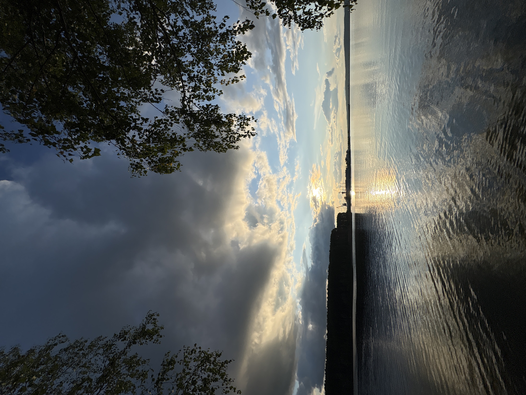 This is a photograph of the Kama River in the city of Chaikovsky, Russia, taken at sunset. The image captures a wide expanse of water with gentle ripples reflecting the sky. In the distance, a dark, tree-covered shoreline is visible, and to the right of the shoreline, industrial cranes and structures suggest a port or industrial area. The sky is a dramatic mix of dark, stormy clouds on the left, transitioning to lighter clouds illuminated by the setting sun on the right. Branches of trees frame the top of the image. The overall mood is tranquil yet moody due to the contrast in the sky. There are no people or animals visible in the scene.