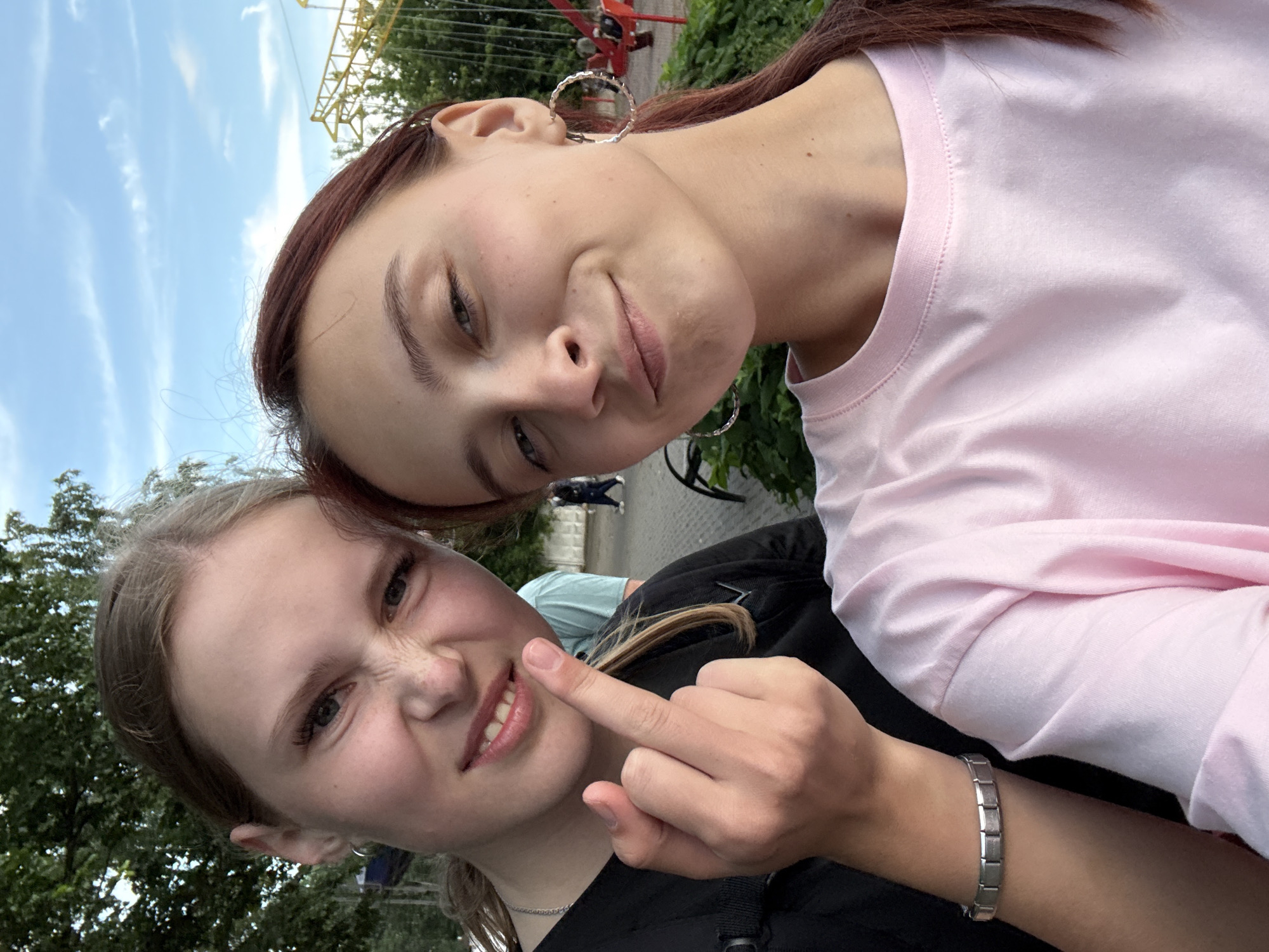 Two young women are taking a selfie outdoors. The woman on the left is making a face and flipping the bird. She is wearing a black shirt and a silver bracelet. The woman on the right is smiling and wearing a pink t-shirt and hoop earrings. In the background, there is a playground with a red swing set, trees, and a cloudy blue sky. The location is likely a public park or recreational area in Izhevsk, Russia, given the presence of playground equipment and typical Russian flora.