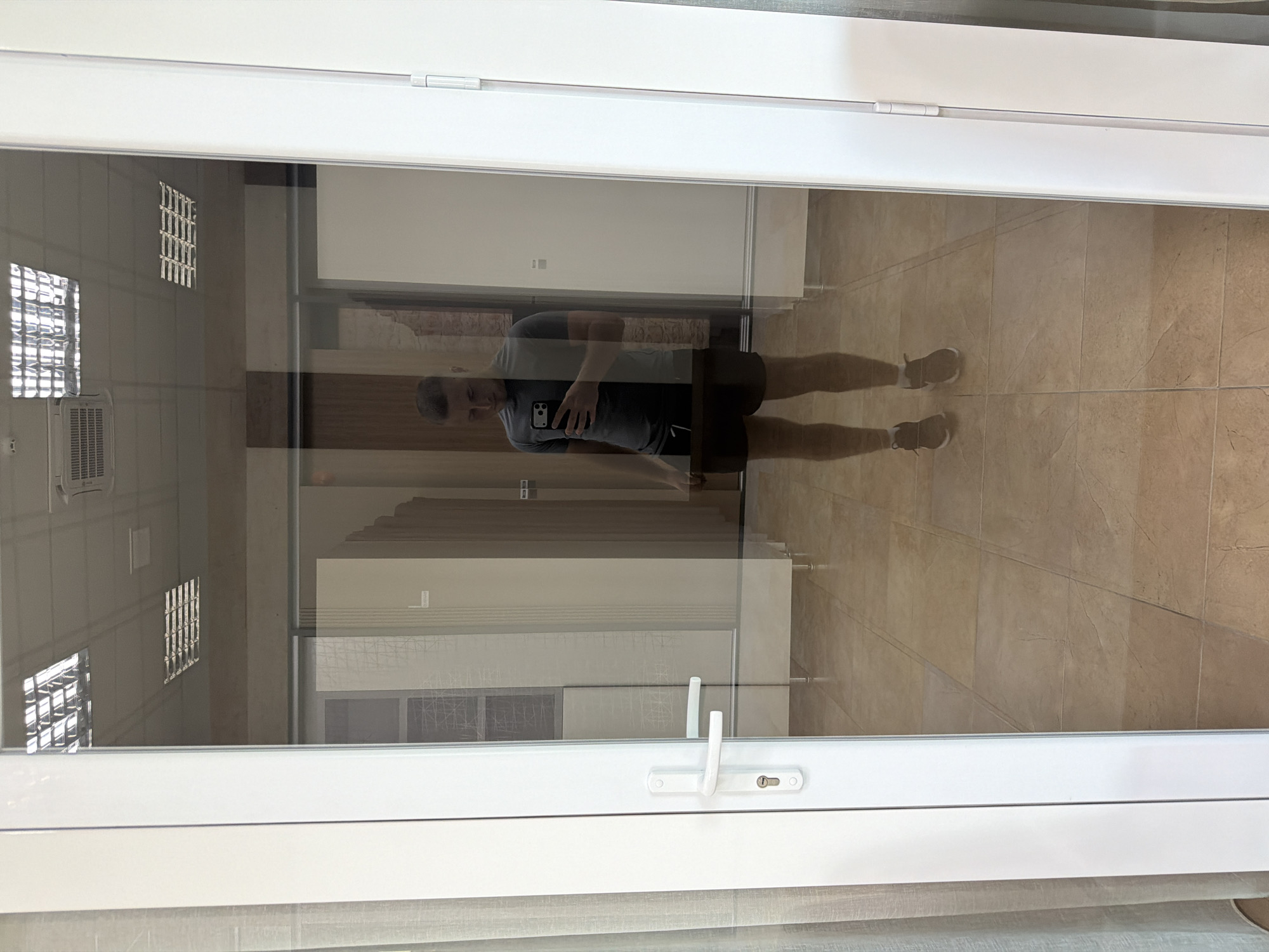The image depicts a man taking a mirror selfie. He is wearing a dark grey t-shirt and dark shorts, with athletic shoes and white socks. The man is standing in what appears to be a store or showroom, indicated by the tiled floor, display walls with various materials, and overhead fluorescent lighting. The reflection also shows a white door with a silver handle and lock on the left side of the frame, and a curtain to the far left. The man is holding a smartphone in his right hand, angled to capture his reflection. The environment appears to be indoors, well-lit, and organized. The photo has been taken in the city of Trușeni, Moldova.