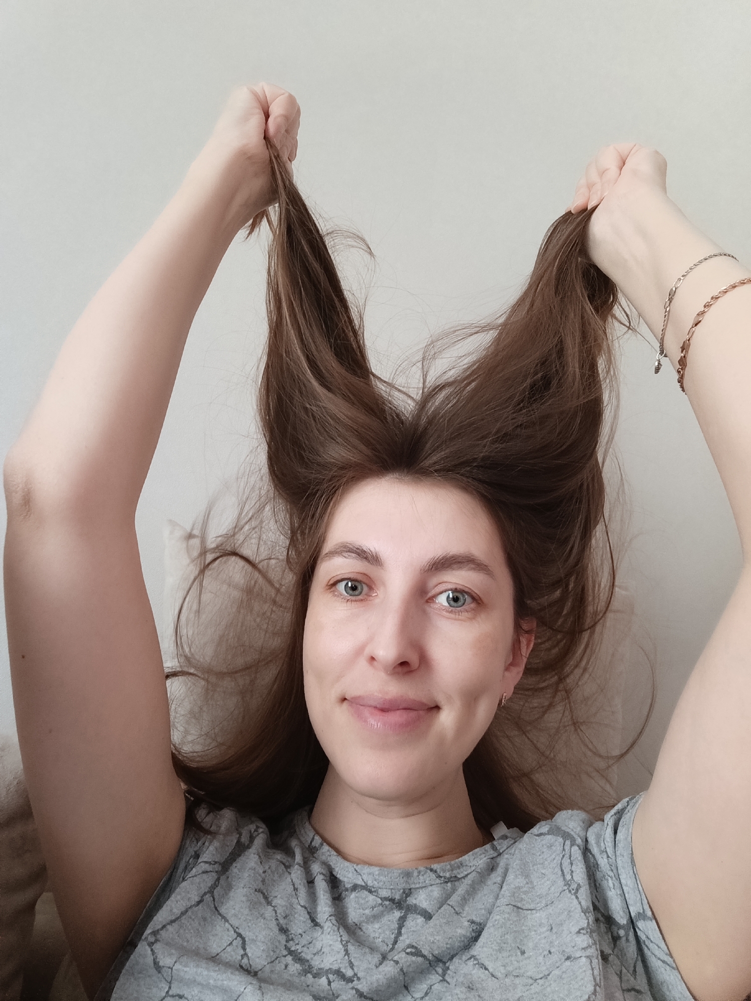 A young adult woman is pictured indoors in Chaikovsky, Russia, looking directly at the camera with a soft smile, conveying a playful and lighthearted mood. She appears to be lying down, with her arms raised above her head. With both hands, she is holding up sections of her long, brown hair, creating two distinct, upward-standing tufts that resemble ears or horns. Her eyes are light-colored, likely blue or green. She is wearing a light gray t-shirt featuring a subtle, abstract, marble-like or crackle pattern in darker gray. On her right wrist, two delicate bracelets are visible: one silver-colored chain and another copper or rose gold-toned braided bracelet with a small, dark charm. The background is a plain, light, neutral wall or surface, suggesting an uncluttered indoor environment. The lighting is bright and even, indicative of daytime, though the specific time of day or outdoor weather cannot be determined. No discernable text is visible in the image.