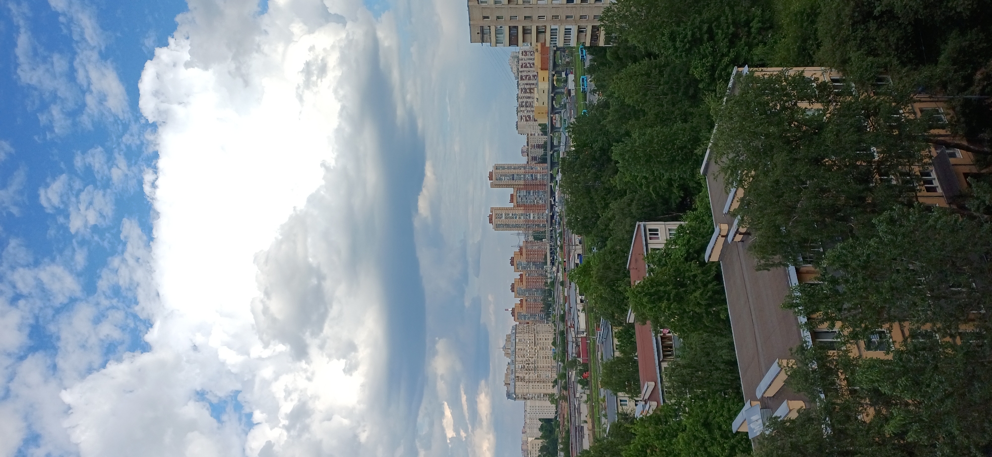 The image captures an urban landscape in Saint Petersburg, Russia, viewed from an elevated perspective. The upper portion is dominated by a bright, partly cloudy sky, featuring large, luminous white clouds alongside patches of clear blue sky with smaller, scattered clouds.
Below the sky, a dense urban environment stretches horizontally, comprising numerous multi-story residential buildings, primarily apartment blocks, showcasing varied architectural styles and colors characteristic of Soviet and post-Soviet development. A street or thoroughfare with a few distant vehicles, possibly buses, is visible amidst the buildings. The urban area is rich with green foliage, with dense clusters of trees interspersed among the structures and along potential streets.
In the immediate foreground, several rooftops are discernible, mostly brown or gray, partially obscured by abundant green trees. A distinct yellow building with multiple windows is prominent in the mid-right foreground, largely surrounded and partially covered by leafy trees.
The scene is brightly lit, indicating it is late morning or early afternoon. The weather is partly cloudy with strong daylight. No discernible people or specific activities are captured, presenting a static overview of the city. There is no visible text in the image.
