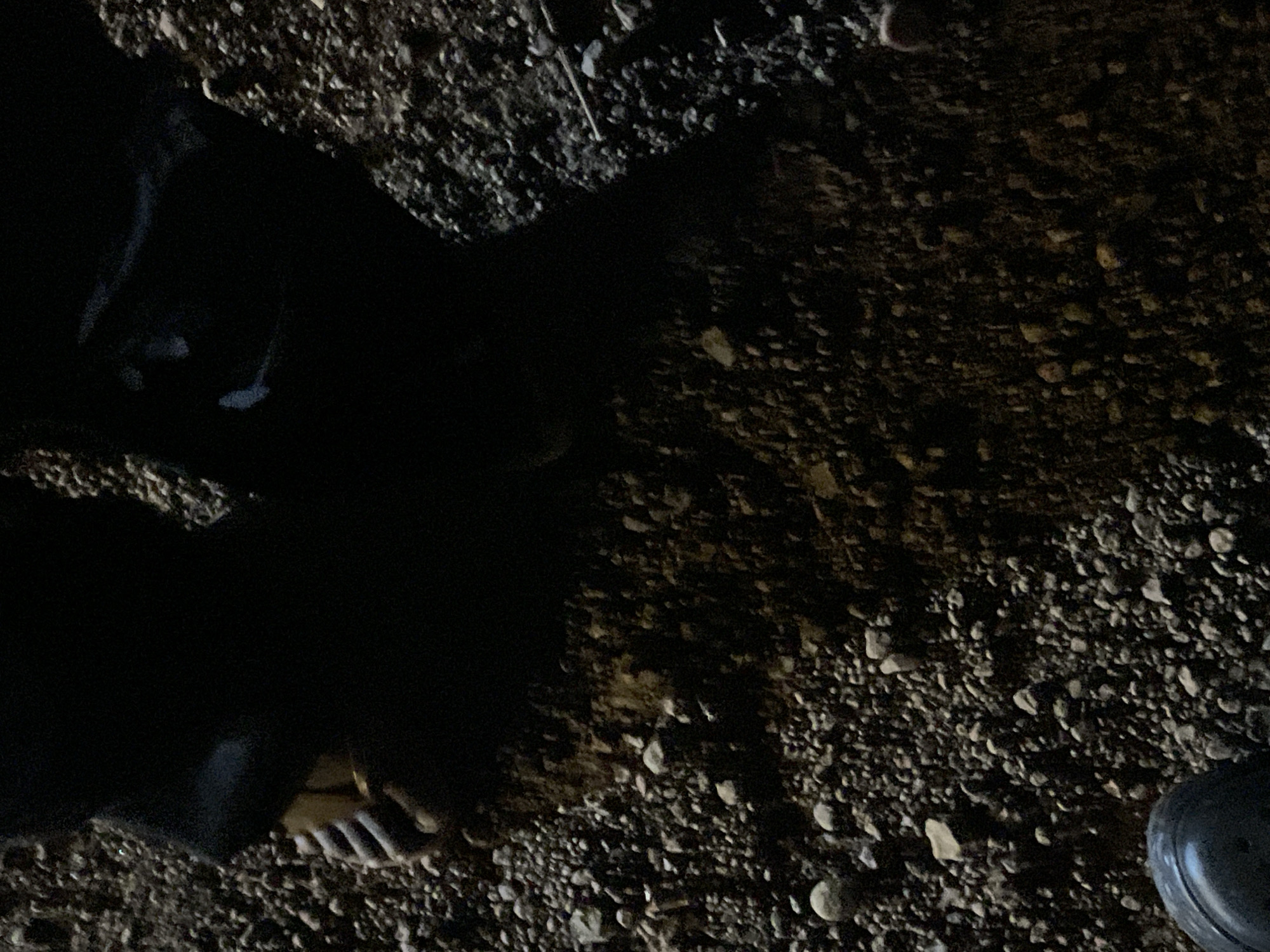 The image shows a close-up view of a person's feet and legs partially submerged in a dark, muddy, and rocky area, likely a ditch or trench. The person is wearing dark clothing and appears to be lying down or squatting. The environment is characterized by loose gravel, dirt, and some larger stones. The lighting is dim, suggesting it might be nighttime or a poorly lit area. There is a dark, round object, possibly a shoe or boot, visible in the upper left corner. The overall scene is gritty and appears to be outdoors in an unpaved area. The location in Durumi, Nigeria is not discernible from the image itself but is provided contextually.