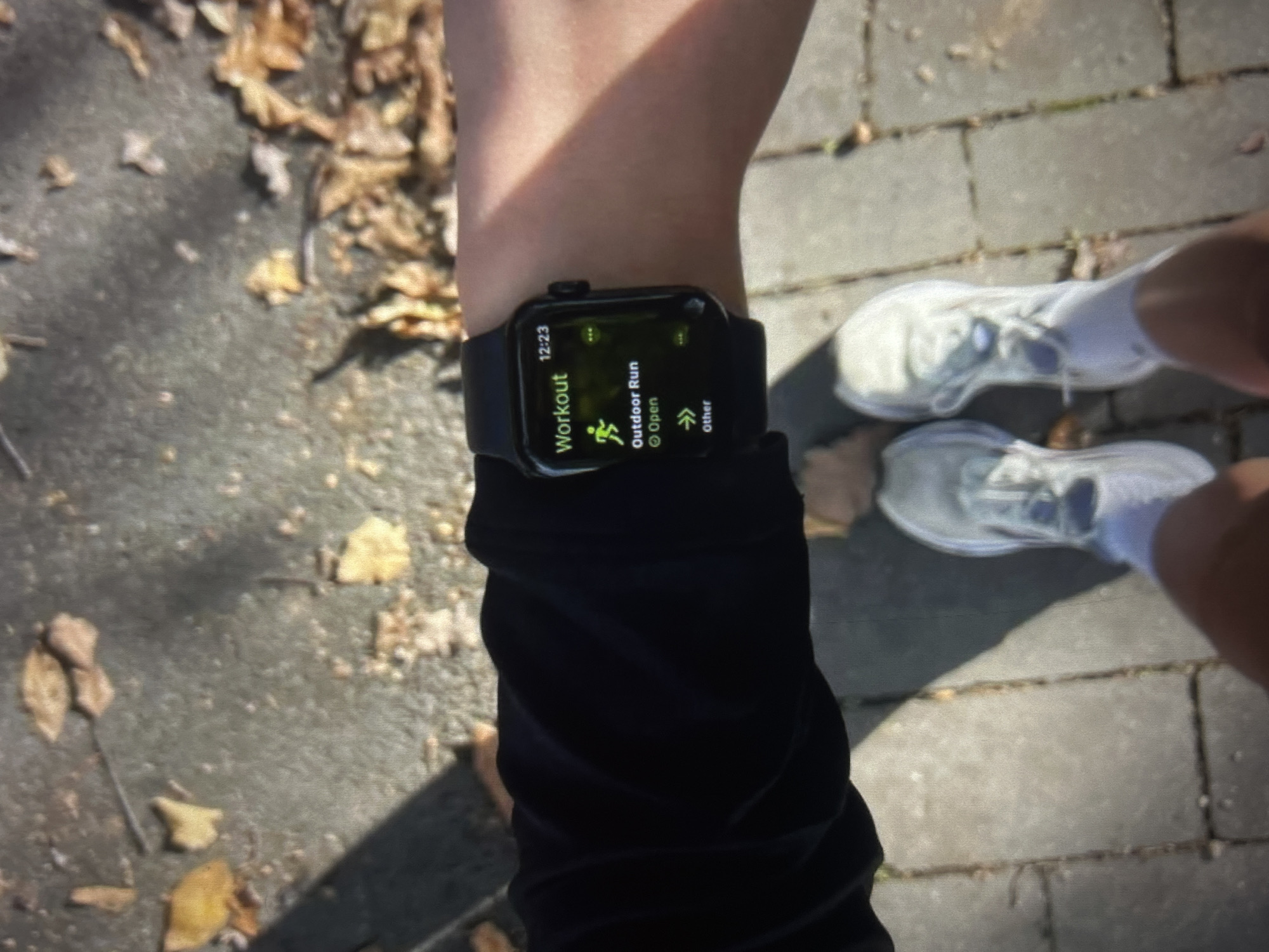 A close-up, low-angle shot shows a person's left arm wearing a black long-sleeved shirt and a black Apple Watch. The watch face displays the "Workout" app, with "Outdoor Run" and "Other" as options. The time is 12:23. The person is standing on a paved walkway with fallen autumn leaves scattered on the ground. Their feet are clad in white sneakers. In the background, out of focus, is a blurred view of more paved ground and autumn foliage. The scene appears to be outdoors on a sunny day.