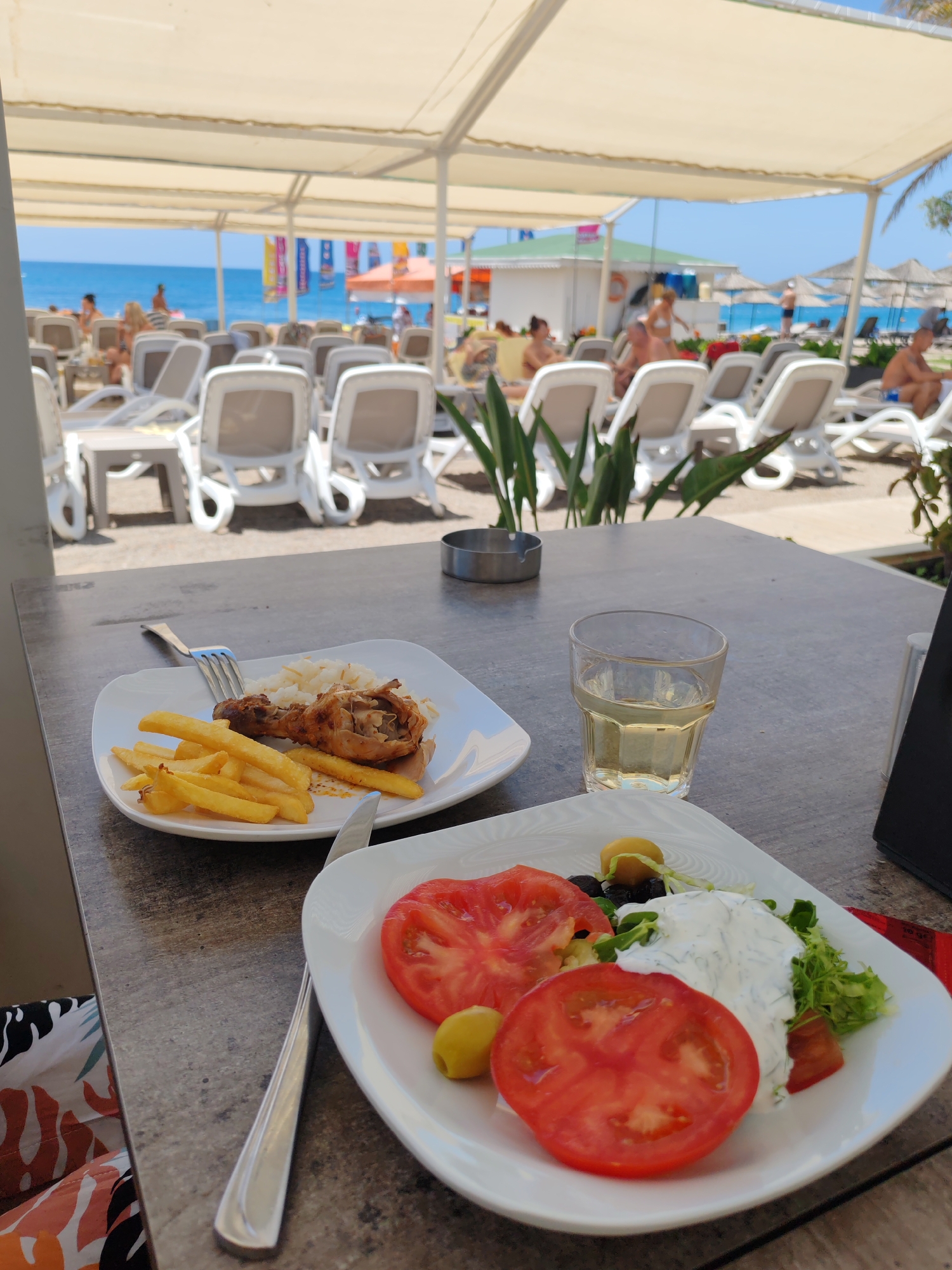 The photograph was taken at an outdoor eatery in Alanya, Turkey. In the foreground, a stone-look table holds two square, white plates of food and a glass of liquid. The plate on the left contains a portion of French fries, rice, and a piece of chicken. A fork and knife rest next to the plate. The right plate features tomato slices, salad, olives, and a dollop of a white, creamy dressing. A glass of a light-colored liquid sits between the two plates. An ashtray is visible behind the glass.
In the background, under a white canopy, are multiple white lounge chairs occupied by people in beach attire, suggesting a resort setting near the beach. The blue sea and sky are visible in the distance. The image is taken during daylight hours.