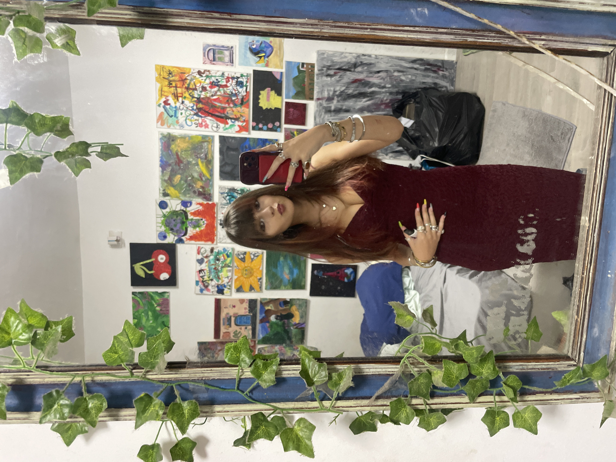 A young woman with long brown hair and bangs is taking a mirror selfie in a bedroom in Raanana, Israel. She is wearing a maroon, form-fitting dress with spaghetti straps and is adorned with several rings and bracelets. She is holding a red smartphone in her right hand, using it to take the photo. The mirror is framed by a rustic, blue-painted wood frame with artificial ivy vines draped around it. The reflection in the mirror shows a wall covered in various paintings, ranging in style and subject matter, including a painting of cherries, a landscape, and abstract art. A bed with a blue comforter and a grey duvet is visible in the background, along with a large black trash bag on the floor and a gray rug. The overall impression is of a creative and eclectic living space.