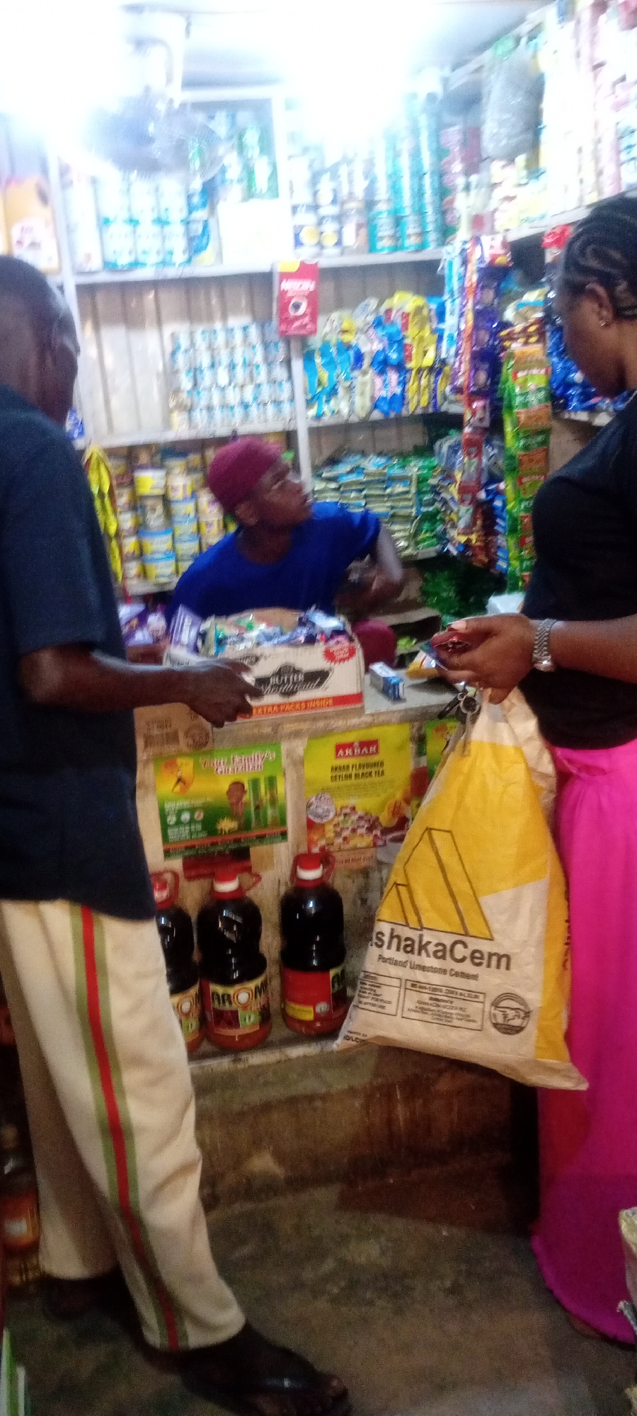 The image shows a scene inside a small store in Kano, Nigeria. A man in a blue shirt and a red head covering is behind the counter serving customers. A man wearing cream-colored pants with green and red stripes on the sides is standing at the counter. A woman in a black shirt and a pink skirt is on the right, holding an item in her hand. A yellow bag labeled "shakaCem Portland Limestone Cement" is on the counter in front of her. Bottles of what appears to be juice are displayed near the counter, labeled "AROM". A box of "BUTTER SHORTBREAD" is at the corner of the counter. "AKBAR Flavoured Ceylon Black Tea" is on the wall. Shelves behind the counter are stocked with various consumer goods. The floor appears to be concrete.