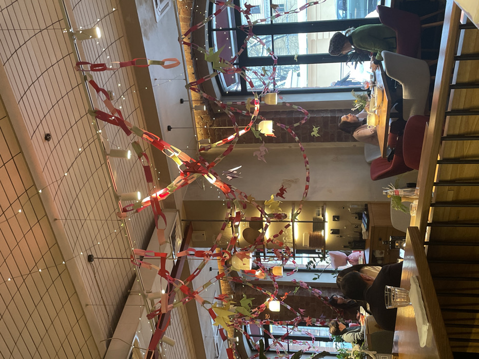This image depicts the interior of a cafe or restaurant in Saint Petersburg, Russia, characterized by its festive decor and relaxed atmosphere.
The scene is dominated by an elaborate network of colorful paper chains and origami figures, primarily red, green, white, and pink, strung horizontally and vertically across the ceiling and around light fixtures. Numerous origami shapes, resembling stylized birds or horses, are integrated into these paper garlands. The ceiling itself features a grid-like pattern with recessed lighting and several modern pendant lights. Additional track lighting elements are visible, illuminating the space.
The interior architecture combines exposed red brick walls with lighter, possibly painted or plastered sections. Large arched windows line one side of the establishment, revealing an outdoor street scene under what appears to be a snowy or cold day, characteristic of Saint Petersburg winters. Through the windows, a traditional European-style building with ornate window frames is visible across the street, along with a dark colored car parked on the side. The exterior environment further confirms the setting in a typical urban area of the city.
Inside, several individuals are seated at tables. In the mid-ground right, two women are engaged in conversation at a light wooden table. One woman, wearing a light top, faces the other, who is dressed in a green jacket and a patterned, possibly knitted, hat. A drink glass and a bowl are on their table. Near the foreground, a wooden counter or railing is visible, with a glass of water and a white napkin placed on it. Further into the cafe, towards the left and center, other patrons are seated at tables; one individual is seen from behind wearing a dark jacket, and others are partially visible, suggesting a moderate occupancy.
A wooden cabinet or bar area against the far left wall features shelves holding bottles and various decorative items, including a pink, wrapped object that resembles flowers. The overall lighting is warm, with light sources providing a soft glow throughout the interior. Actions observed include dining, drinking, and social interaction. A specific street address for the location is not discernible from the visual information provided within the image, but the architectural and environmental cues are consistent with central Saint Petersburg.