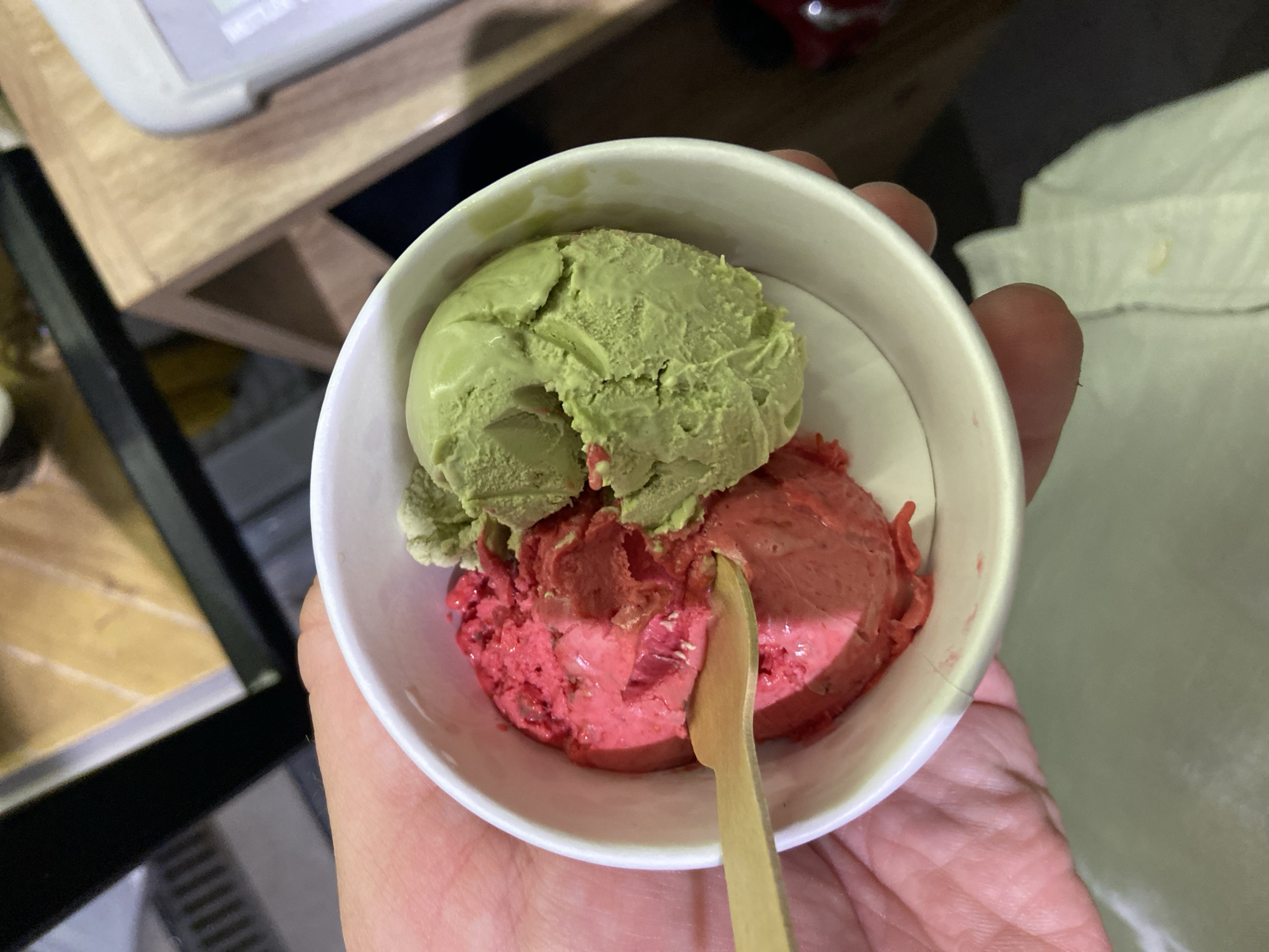 A person holds a white paper cup containing two scoops of ice cream: one green and one red. A small wooden spoon is partially inserted into the red scoop. The person's hand is visible in the foreground, and a white shirt is partially visible on the right. The background includes a wooden table with a white object on top, and what appears to be a black metal frame. The location is suggested to be Petrovac centar, Montenegro.