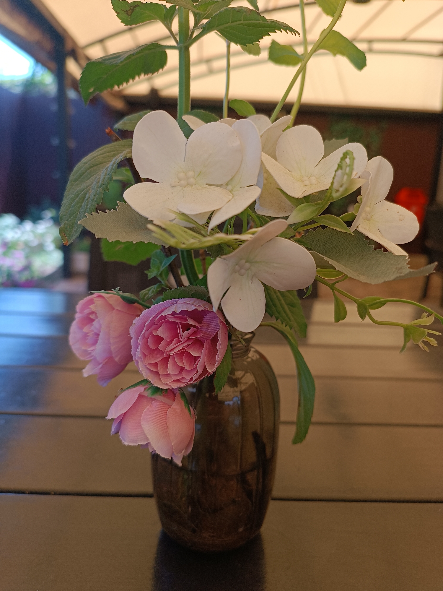 A vase containing pink roses and white flowers sits on a dark wooden table outdoors. The vase is dark brown glass and is partially filled with water. The roses are a vibrant pink, and the white flowers appear to be hydrangeas. The table is made of dark wood planks. In the background, a light-colored awning is visible, as well as some greenery and a blurry red object. The scene is brightly lit, suggesting it is daytime. The location is a patio or outdoor seating area, possibly in a garden or restaurant. There are no people visible in the image.