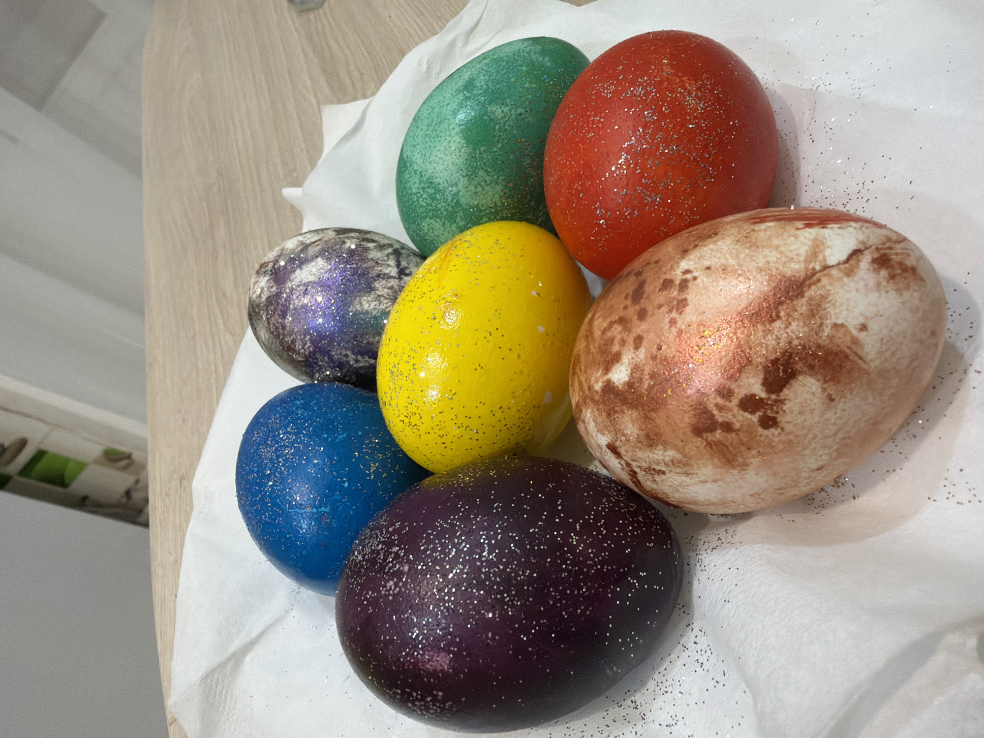 The image shows seven decorated Easter eggs resting on a crumpled white cloth on a wooden surface. The eggs are colored in various shades including green, red, purple, yellow, blue, and a mottled brown and white. Glitter has been applied to all the eggs. The location is not indicated in the photograph.