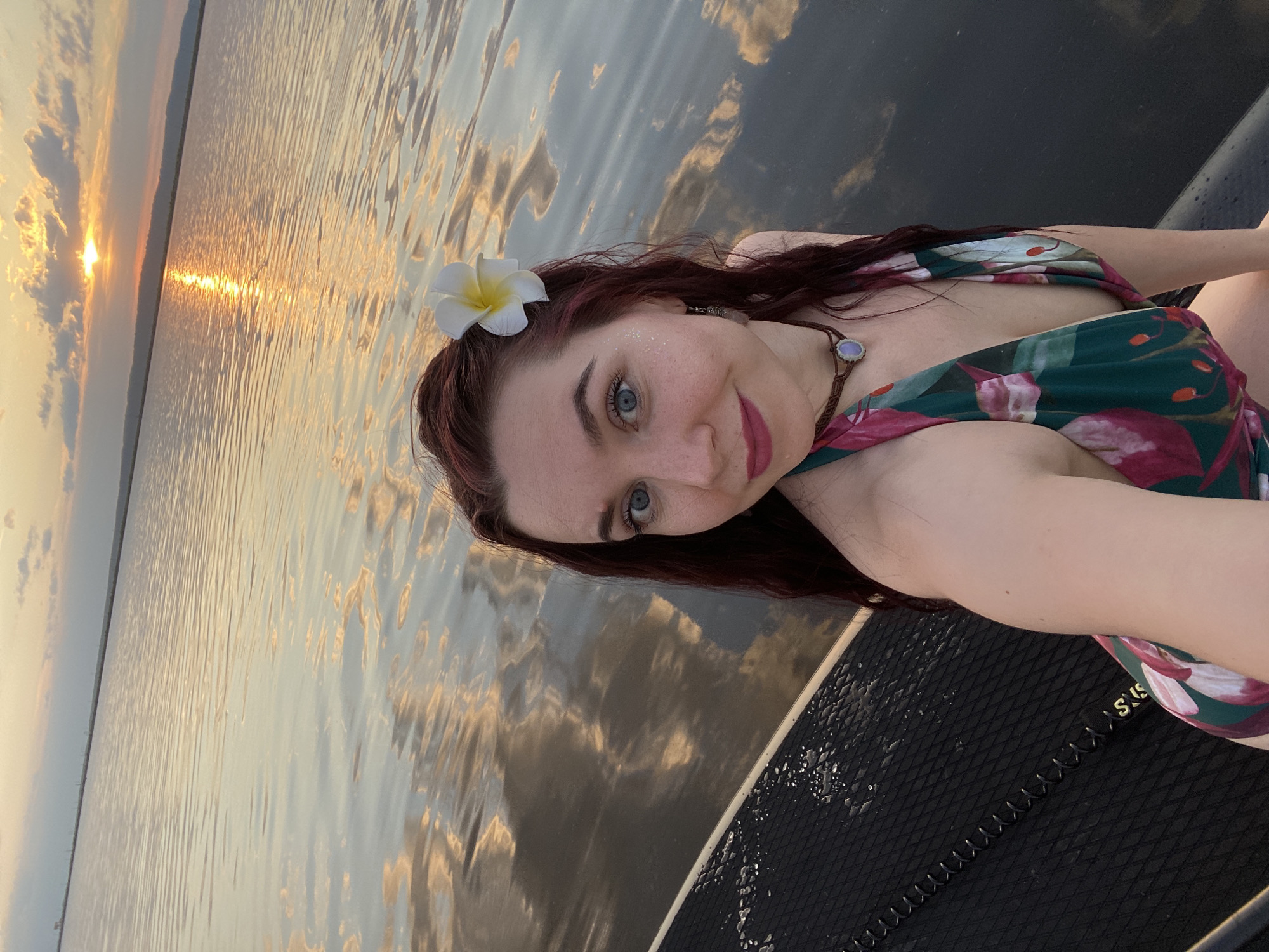 A selfie shows a woman on a black paddleboard on a body of water. The woman has fair skin, red hair adorned with a white flower, and blue eyes. She is wearing a patterned swimsuit with green, pink, and white hues. She is looking directly at the camera with a slight smile. The background features a reflective surface of water, with a sunset visible on the horizon. The location is likely in the city of Chaikovsky, Russia.
