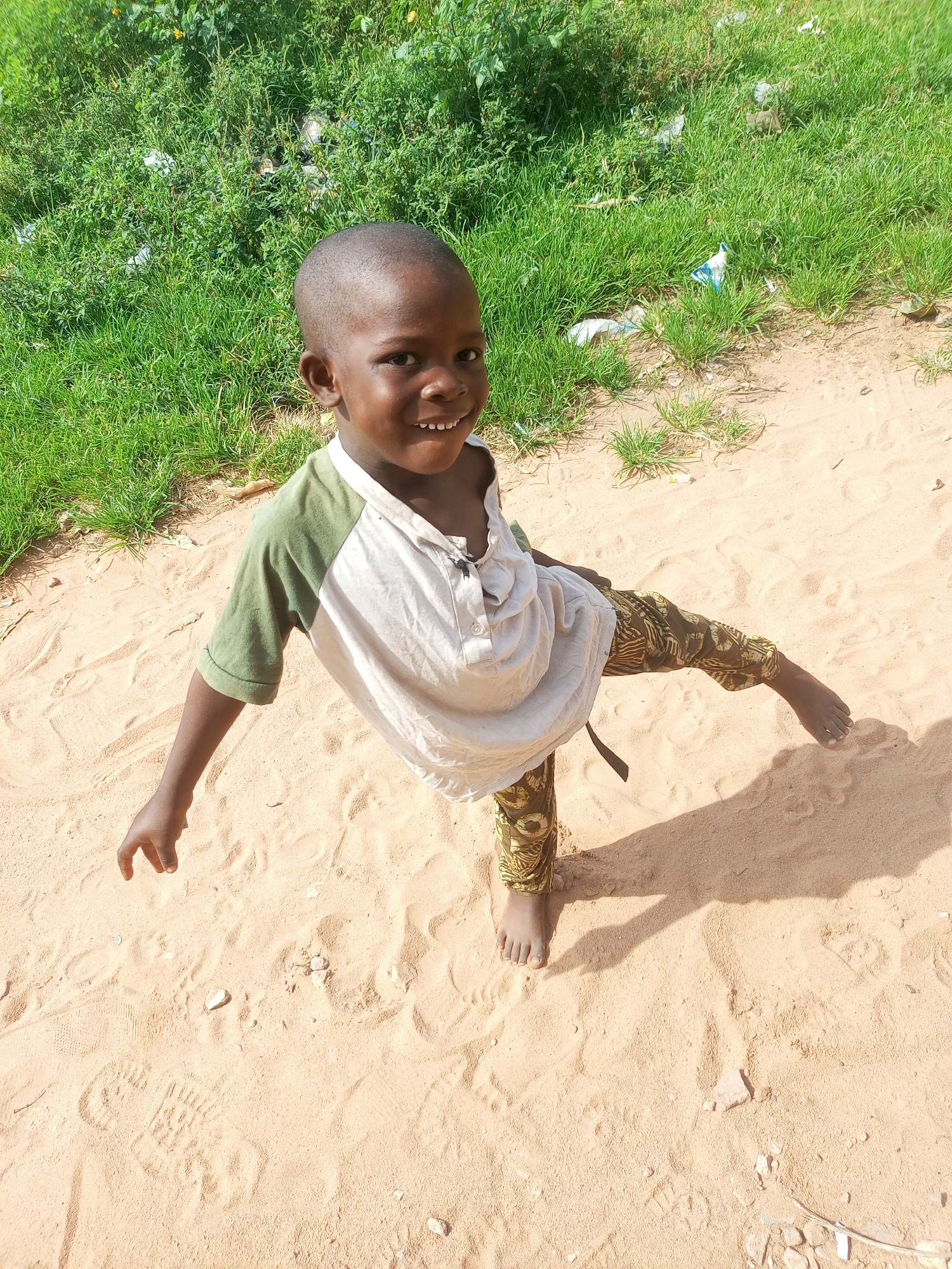 The image depicts a young boy standing on sandy ground, with a patch of green vegetation in the background. The boy is looking towards the camera with a smile, showing his teeth. He is wearing a short-sleeved shirt with a green collar and sleeves, and patterned pants. His left leg is extended forward. The ground is sandy with some footprints and small stones visible. The vegetation appears somewhat overgrown and includes some litter. The lighting suggests it is daytime. The location is specified as Tempure, Nigeria.