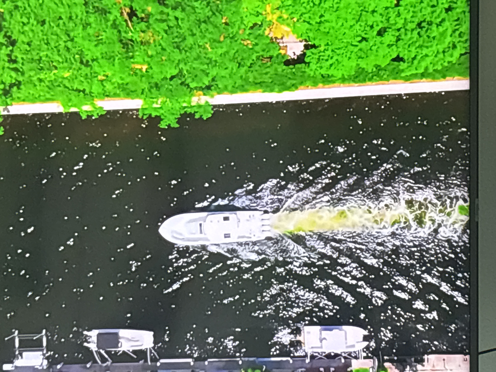 This is an aerial, over-the-shoulder shot of a white motorboat traveling through dark, choppy water, leaving a foamy wake. The boat is heading away from the viewer, and its stern is emitting a bright yellow-green exhaust. To the right of the boat, the water is bordered by a bright green, dense tree line. To the left of the boat, there is a dock with several boats moored, and the water is darker with more visible reflections. The scene is likely a canal or waterway in a developed area, possibly within Dubai, UAE, given the presence of a modern dock and lush greenery.