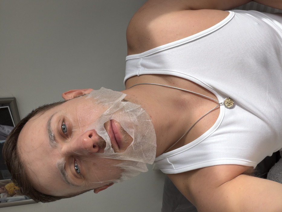 A male individual, appearing mid-adult, is depicted in a close-up, chest-up indoor shot within a room in the city of London, United Kingdom. He has short brown hair and blue eyes, and is looking directly towards the camera. A white sheet face mask is applied to his face, covering the forehead, cheeks, and chin, with cutouts for the eyes. The mask's mouth area is visibly peeled down below his nose, revealing his lips. He wears a white ribbed tank top. Around his neck is a silver chain necklace with a circular, two-tone (silver and gold) pendant. The background features a plain light grey wall, with a partially visible framed picture in the upper left corner showing a light-colored image. Soft, ambient natural lighting illuminates the scene from the left.