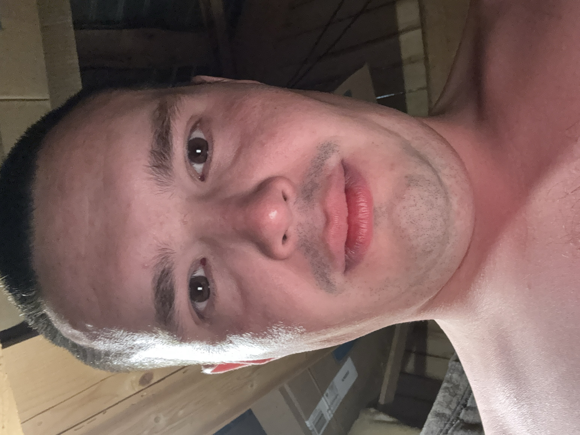 The image is a close-up, front-facing selfie of a person's face and upper chest, taken from a low angle. The subject has short, dark hair, brown eyes, and a faint mustache. Their skin appears flushed. The background consists of wooden beams and what appear to be cardboard boxes, suggesting an indoor, possibly unfinished, space. No specific location in Sharkan, Russia can be determined from the visual information presented.
