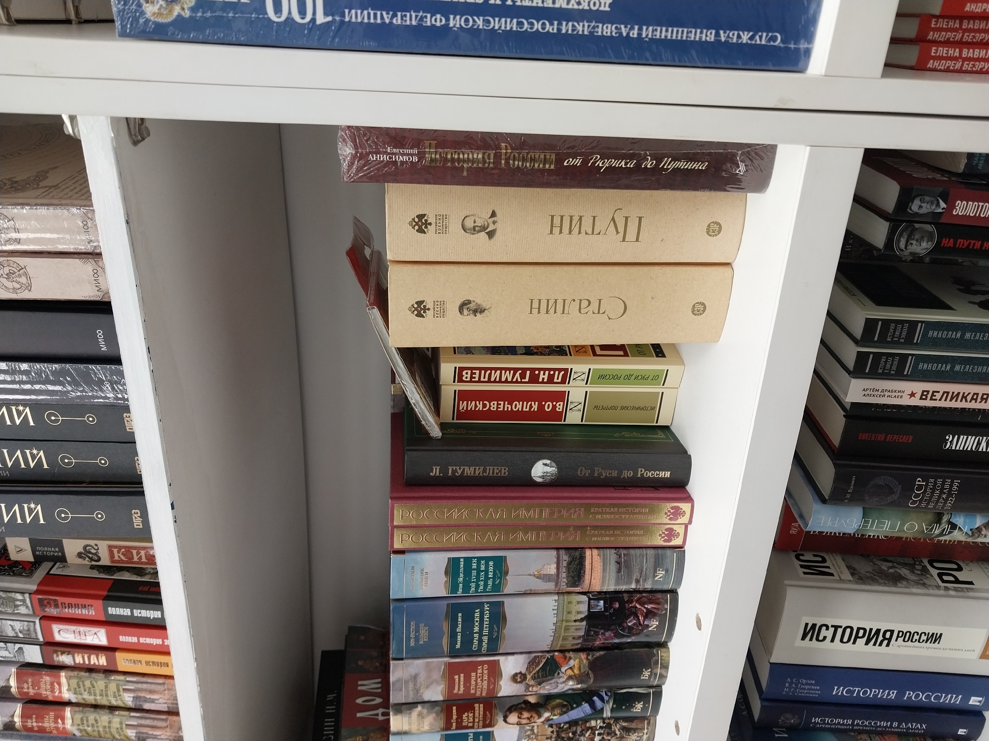 The image shows a shelf filled with books, likely in a bookstore or a private collection. The books are primarily in Russian, with titles covering Russian history and notable figures. The titles include "Putin," "Stalin," and "History of Russia from Rurik to Putin." Other books cover Russian imperial history and the history of Saint Petersburg and Moscow. The shelf is white and appears to be part of a larger bookcase. The image contains books related to Russia and Russian history. It is not possible to determine if this is located in Sochi, Russia.