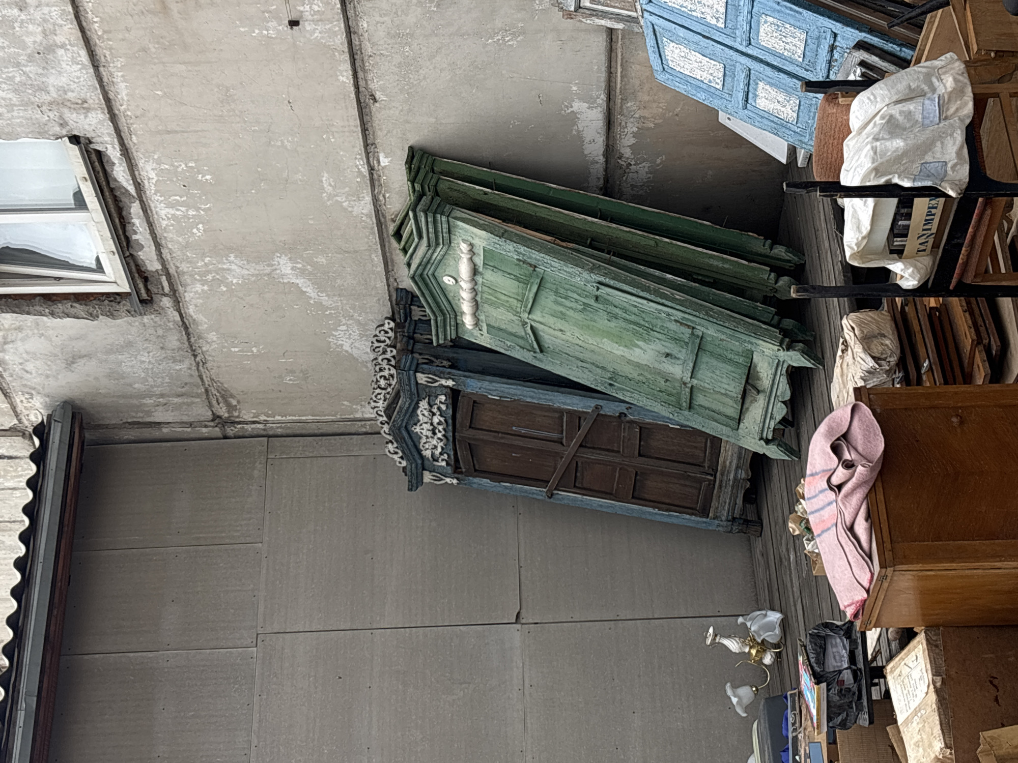 The image appears to be taken in a storage area or warehouse in Krasnoyarsk, Russia. The location contains a variety of objects including several old doors of various colors (blue, green, and brown), some stacked on top of each other. There's also furniture and other miscellaneous items, some covered with cloths or blankets, stored near the doors. A window is visible on a wall in the upper left of the image. The location may be an antique store or a storage space.