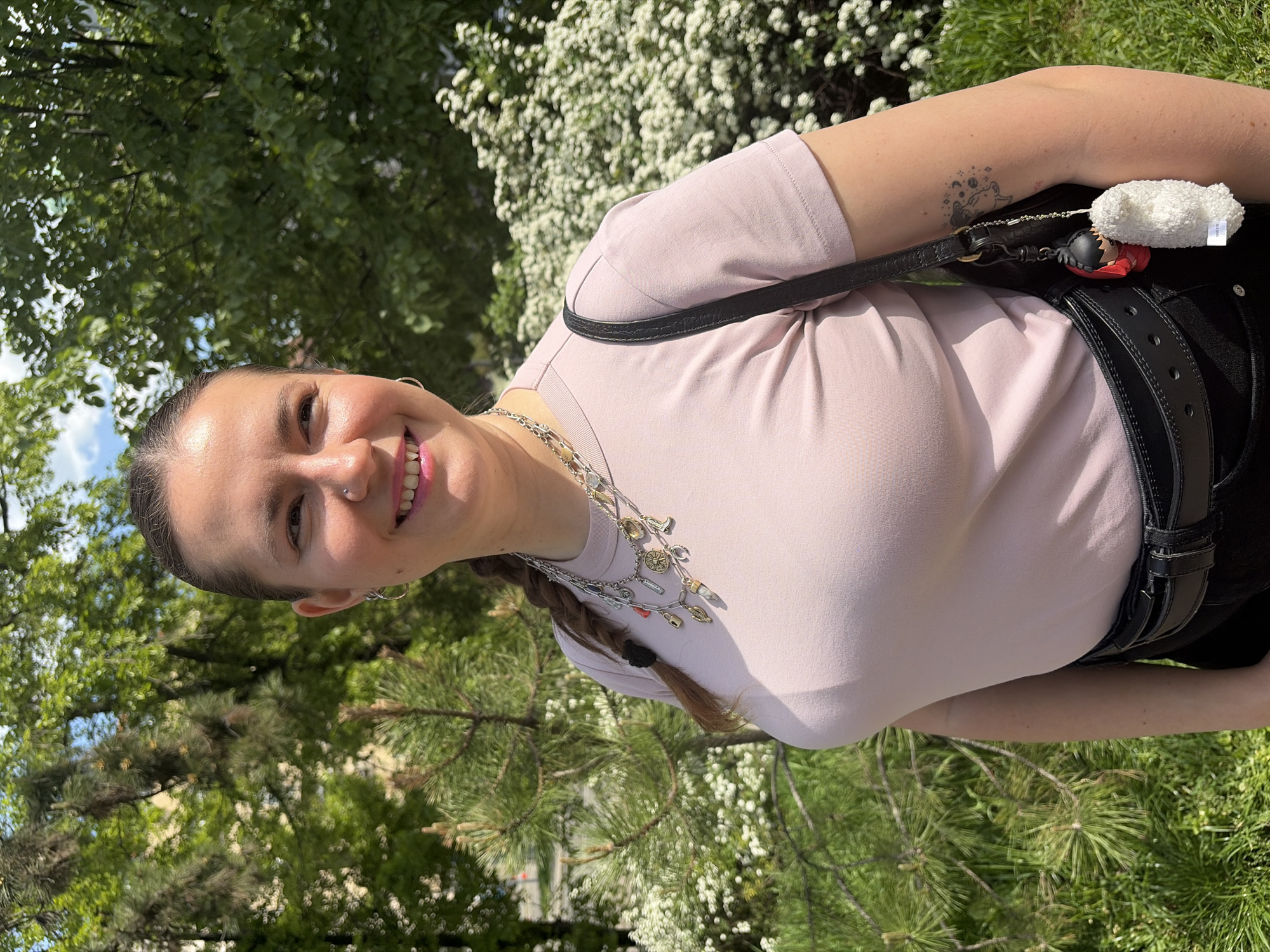 A woman with a single braid and a collection of charms around her neck is standing outside. She is wearing a light pink t-shirt and a black belt. She has a small tattoo on her left arm. She is carrying a black purse with a white and red accessory attached. The background includes various green trees and a flowering bush. The image was taken in Belgrade, Serbia.