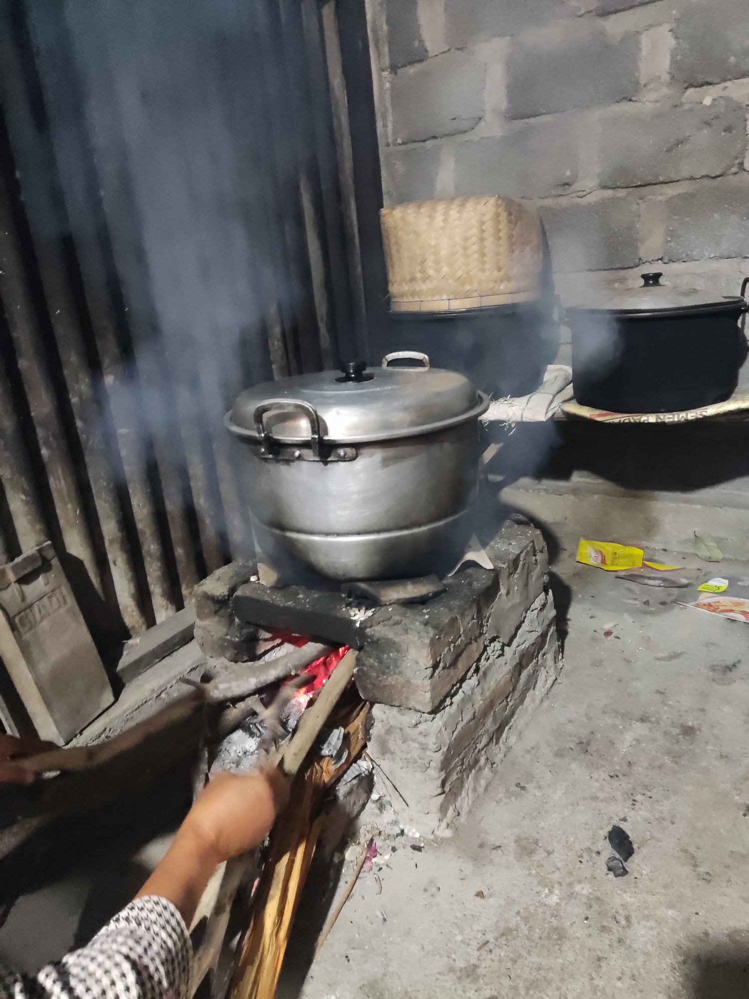 The image depicts a rustic outdoor kitchen scene, likely in Ngentrong, Indonesia, characterized by a traditional wood-fired stove. A large, metallic steamer pot, filled with food and emitting steam, is actively cooking over a fire. Adjacent to it, two dark, lidded pots are also present, one with a woven basket lid. The stove itself is constructed from bricks, with visible glowing embers beneath the steamer. A person's arm and hand, wearing a checkered sleeve, are visible on the left side of the frame, tending to the fire with sticks. The environment appears to be a simple, possibly unpaved area, with a corrugated metal wall in the background and scattered debris on the ground, including packets and what appears to be a leaf. The overall atmosphere suggests a traditional and practical approach to cooking.