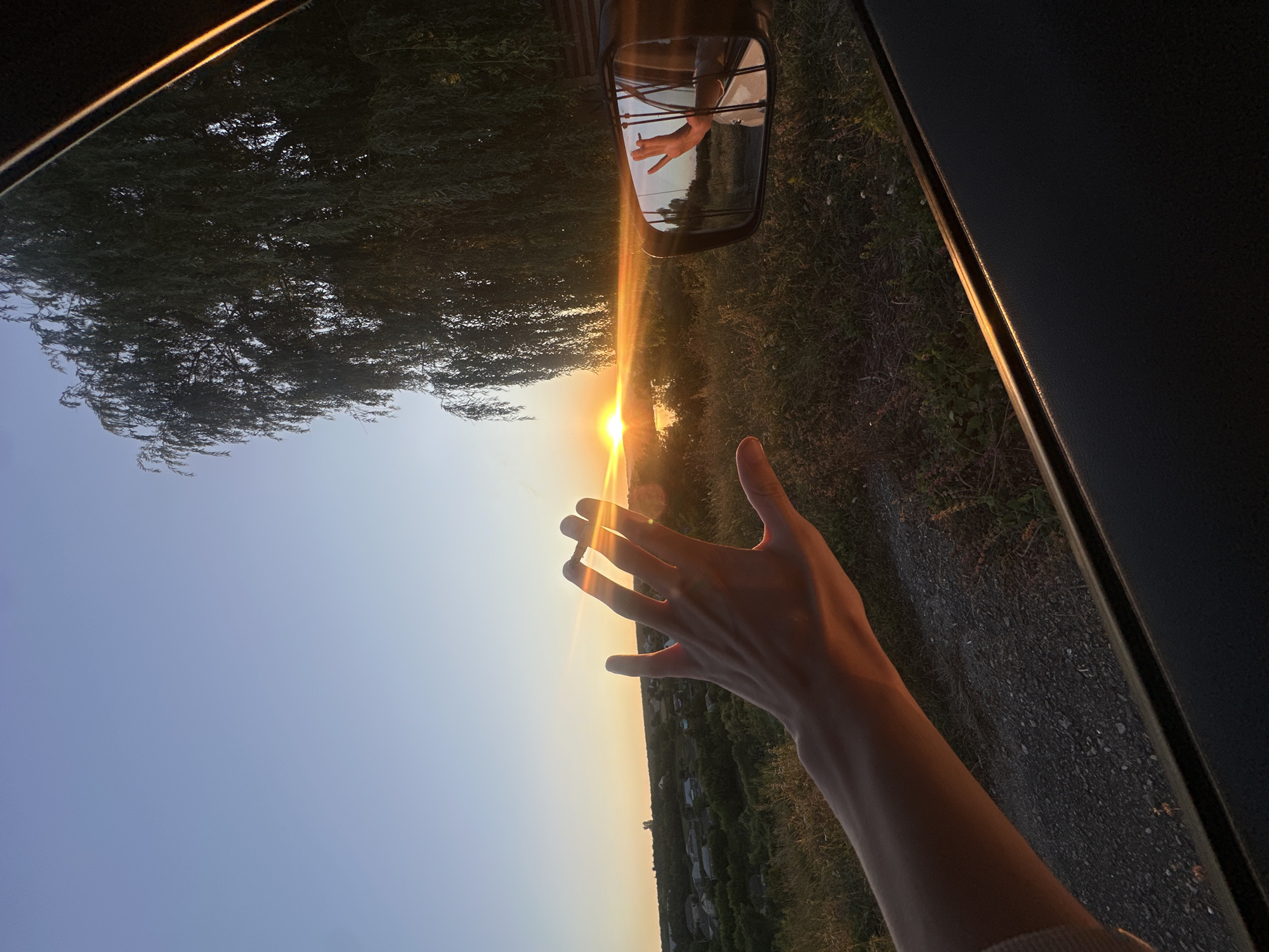 The image captures a sunset scene from inside a vehicle. A person's hand is extended out of the window, reaching towards the setting sun, which is visible over a body of water and a distant village. The sun's golden rays create a lens flare effect. A large weeping willow tree with long, drooping branches is on the right side of the frame, partially obscuring the sky. A car's side mirror reflects the hand and a segment of the sunset. The landscape outside the car consists of grassy fields and a gravel road. The sky is a gradient of clear blue at the top, transitioning to orange and yellow near the horizon. The location is identified as Makiv, Ukraine.