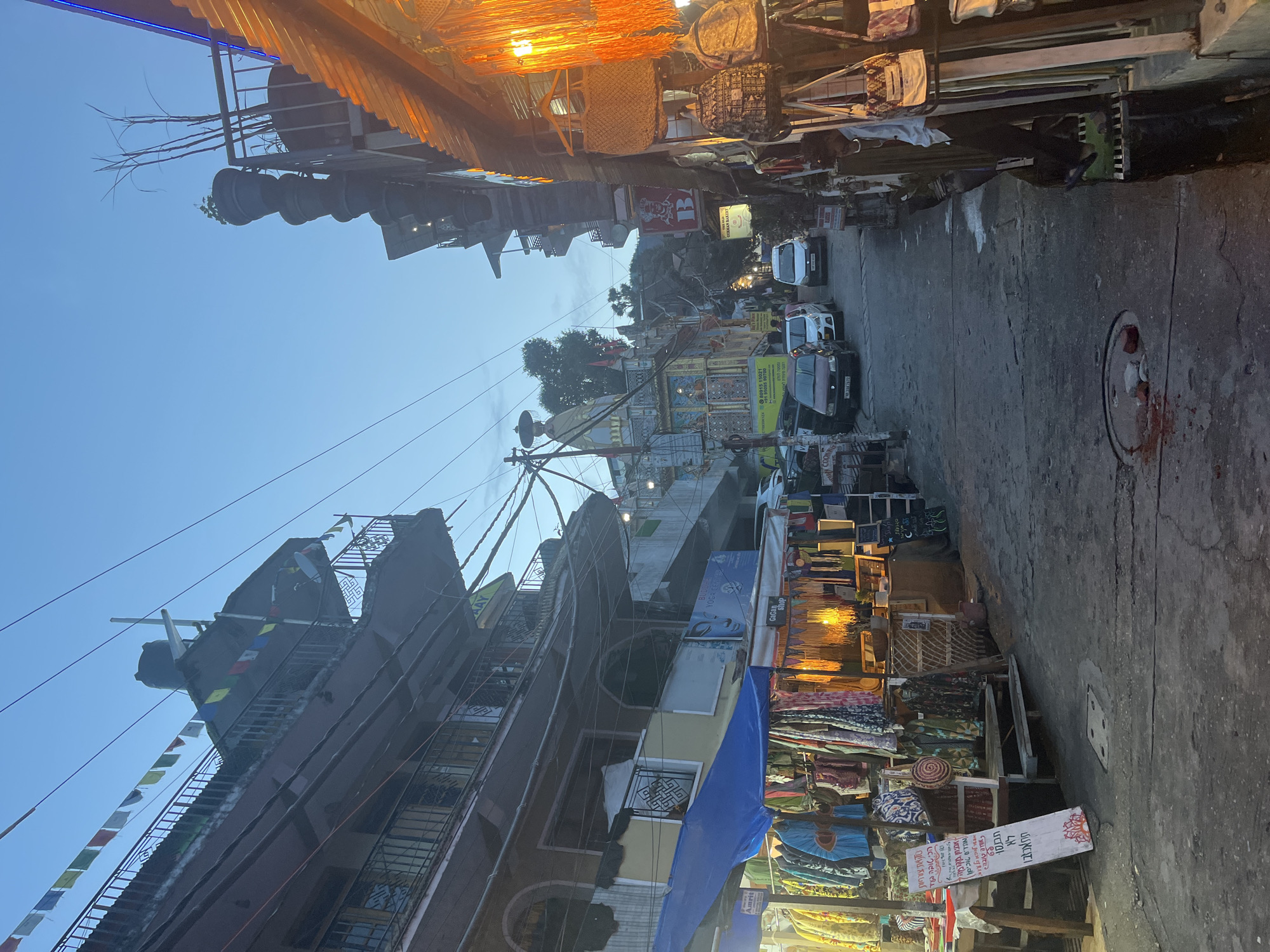 The image depicts a narrow street lined with shops and buildings, likely in a market area, at dusk. The sky is a pale blue, indicating either early morning or late evening. On the left, a multi-story building with balconies and windows is visible. Below it, a market stall is set up with a blue tarp and an array of colorful clothing and textiles hanging for display. A sign with writing in Hebrew is placed in front of the stall.
The street itself is paved and appears to be made of asphalt or concrete, with some visible manhole covers. Several cars are parked along the street, suggesting it's a thoroughfare or accessible by vehicle. On the right side of the street, more market stalls are present, featuring hanging bags and textiles, illuminated by warm artificial light. Prayer flags are strung across some of the buildings.
Further down the street, a prominent structure with a spire, possibly a temple or religious building, is visible, adorned with intricate designs and statues. The overall atmosphere is that of a bustling marketplace, transitioning from daylight to artificial illumination. No specific people are clearly identifiable or engaged in significant actions, though several figures can be vaguely seen in the background. The environment suggests a location with a blend of commercial activity and cultural elements, likely in a region with significant Buddhist or Hindu influence, given the prayer flags and temple-like structure.