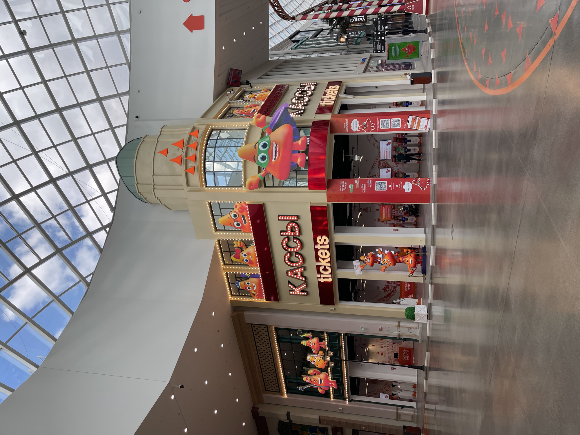 The image shows the interior of a large, modern shopping mall with a glass ceiling. In the foreground, the polished floor reflects the ceiling and the structures above. To the right, there is a curved pathway marked with red triangular patterns. In the center of the image, a brightly colored, cartoon-like structure resembling a building facade stands out. This structure features large, illuminated signs that read "КАССЫ" (KASSY) and "tickets" in English. There are animated cartoon characters with cone-shaped heads and smiling faces displayed on and around this facade. One large character, resembling a superhero with a cape, is prominently featured in front of the "КАССЫ" sign. To the left of this central structure, another section of the mall features a storefront with similar cartoon characters displayed in windows. The overall impression is a lively, entertainment-themed area within the mall.
