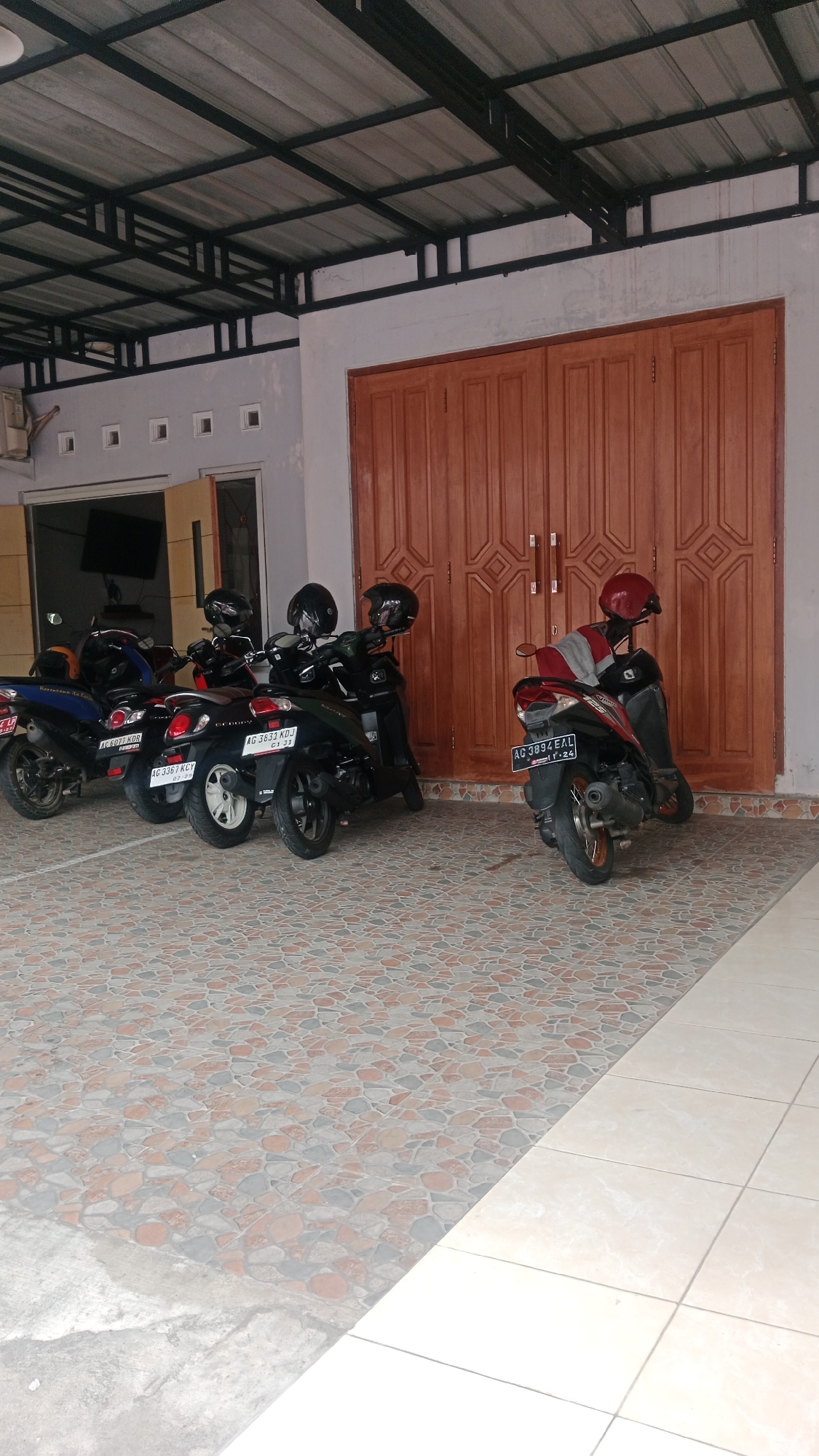 This image captures a covered outdoor area in the city of Blitar, Indonesia, likely a parking space or a veranda. The scene features several motorcycles parked in a row, some with helmets placed on their seats. The motorcycles are of various colors, including blue, black, and dark green. They are parked on a patterned tiled floor. Behind the motorcycles, a large wooden double door is visible, with ornate carvings. Above the door and motorcycles, a metal roof structure supported by beams is present. To the left of the door, a portion of a building wall with several small square windows can be seen, along with an air conditioning unit. The overall impression is one of a functional, residential, or commercial space in Indonesia.