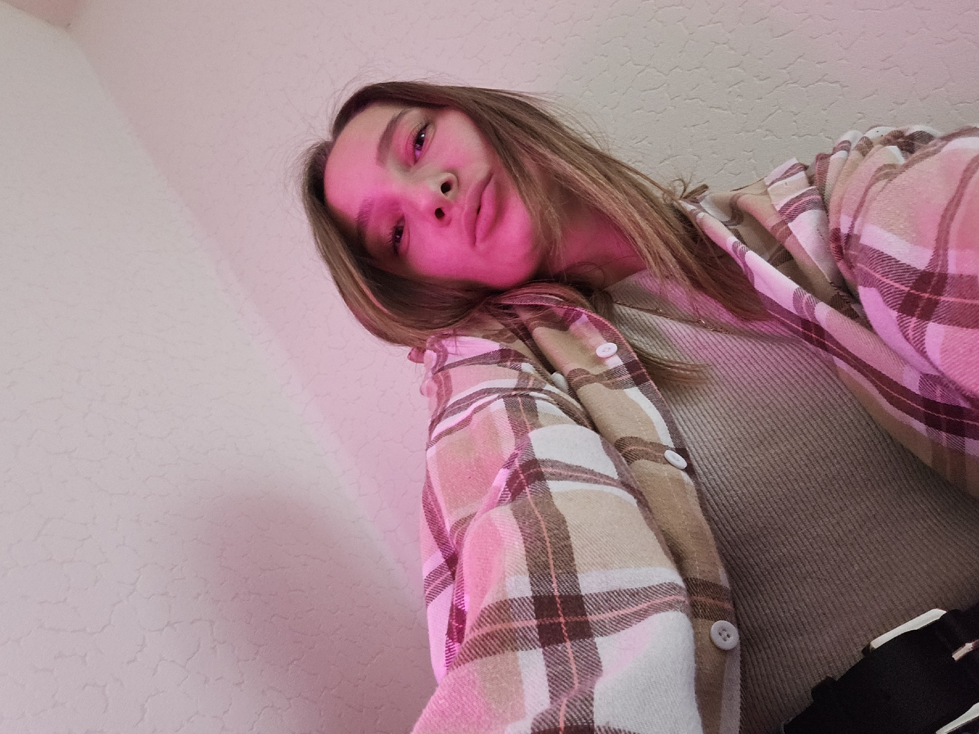 A young female, likely a teenager or young adult, is depicted in a low-angle close-up shot, gazing directly into the camera. Her head is tilted to her right, and her lips are pursed in a slight pout. Her long, light brown hair falls over her shoulders. A dominant, strong magenta-pink light illuminates her face and upper body, casting a pink glow on her features and the surrounding environment. She is wearing a plaid button-up shirt in a pattern of beige, light pink, white, and dark brown squares, layered over a ribbed, light brown or beige top. A thin, gold-colored chain necklace is visible against the ribbed fabric. A small segment of black fabric with a white stripe is visible at the very bottom right, possibly part of lower attire. The background is a plain, light-colored wall (off-white or pale beige) with a subtle, irregular textured pattern. The strong pink light from the subject also reflects onto the upper right portion of the wall, creating a soft gradient. There are no discernible objects, landmarks, or identifying markers in the image that indicate the location as the city of Chaikovsky, Russia.