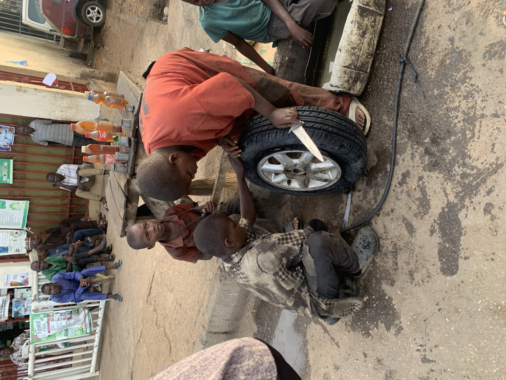 In Gura Top, Nigeria, a group of boys are shown interacting with a car tire. One boy, wearing an orange shirt, holds a large knife and appears to be working on the tire. Two other boys are positioned near him, with one sitting on the ground. A black hose is connected to the tire. In the background, there are people sitting or standing near a building with advertisements or posters. Bottles containing a yellow liquid are placed on a table. A red car is partially visible in the top left corner. The scene takes place outdoors on a concrete or paved surface.