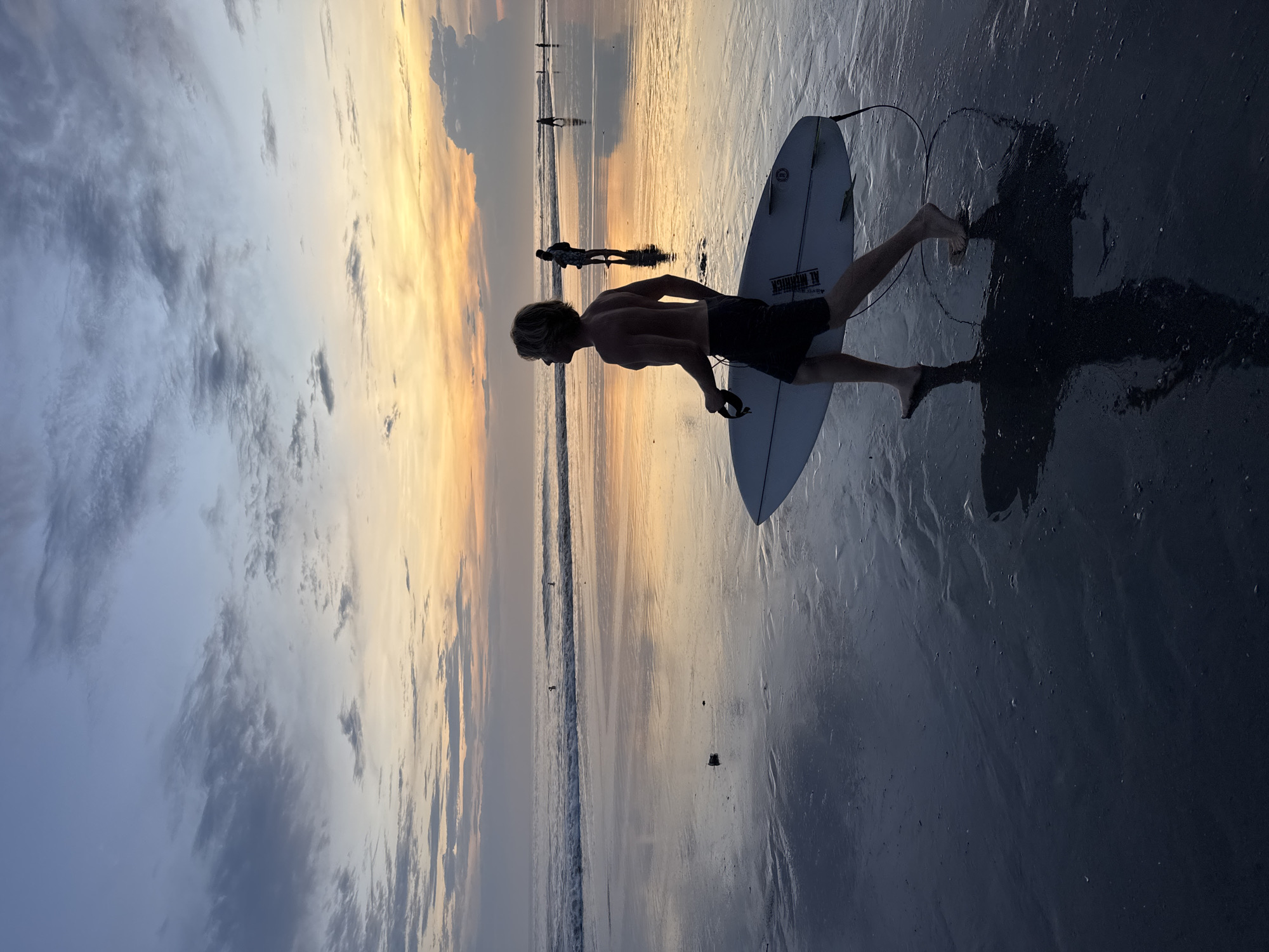The image depicts a beach at sunset in North Kuta, Indonesia. A shirtless man with blond hair is walking on the wet sand, carrying a white surfboard under his left arm. The sky displays a gradient of colors from blue and grey clouds to orange and yellow hues near the horizon. The wet sand reflects the sky, creating a mirror effect. In the distance, there are other figures walking along the shoreline. The man and his surfboard cast a shadow on the wet sand. The surfboard has the inscription "Al Merrick" on it.