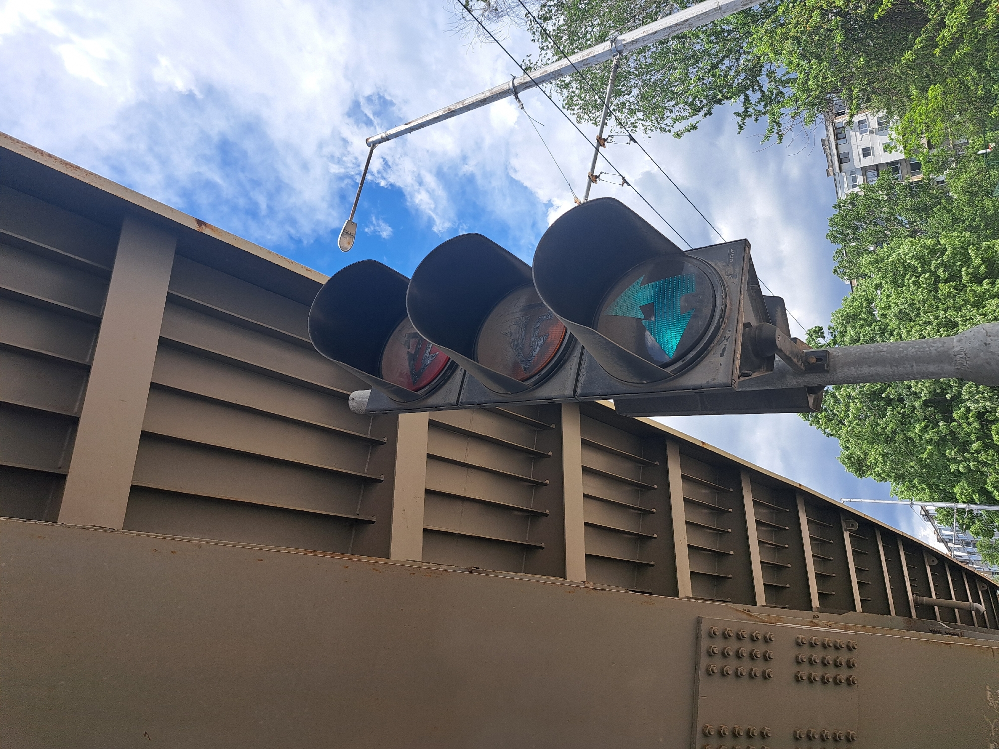 The image shows a traffic light with a green left turn arrow illuminated, mounted on a gray metal pole. The pole is attached to a light gray steel bridge or overpass structure. The structure is heavily bolted together. Above the structure and traffic light, there are utility lines against a backdrop of a partly cloudy sky and the tops of green trees. A white apartment building is visible behind the trees. While difficult to confirm with certainty, the general style of the buildings and infrastructure, and the specific type of traffic light used suggests this photo was taken in Sofia, Bulgaria, although further information such as street signs would be needed to pinpoint the exact location. There are no people visible in the image.
