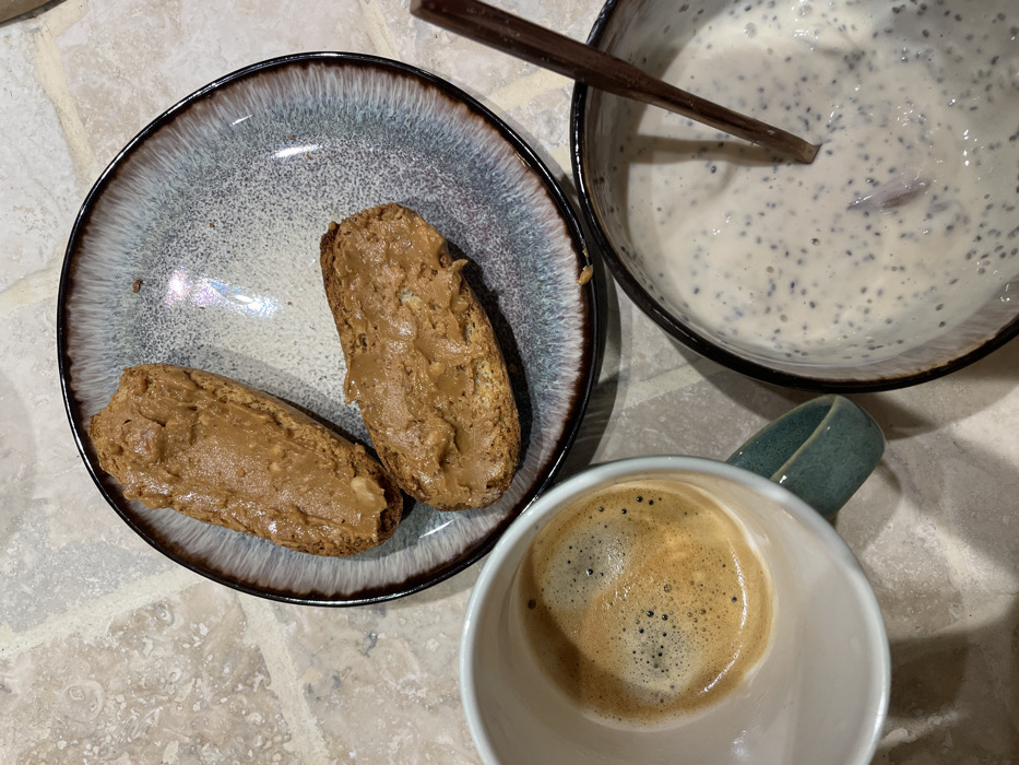A top-down shot reveals a nutritious breakfast spread. Two slices of toast lavishly spread with peanut butter sit on a speckled blue plate with a dark rim. Adjacent is a bowl of chia seed pudding, featuring a wooden spoon resting inside. A white mug filled with frothy espresso is placed next to the bowl, completing the meal arrangement. The breakfast items are set against a textured, neutral-toned surface.