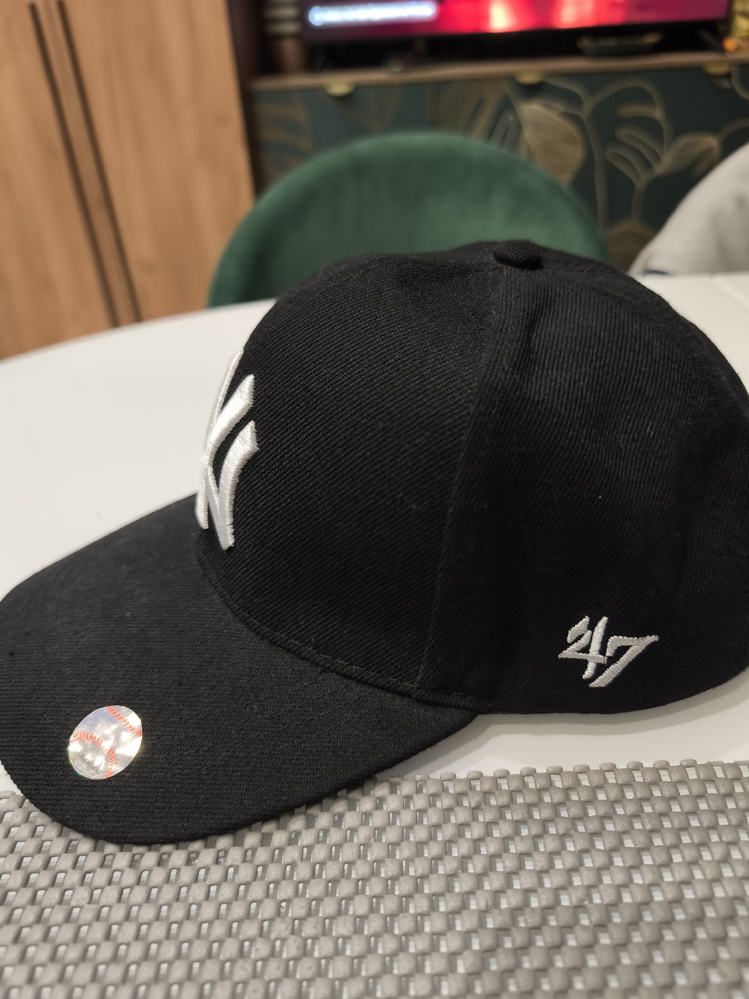A black New York Yankees baseball cap sits on a gray, textured surface. The cap features a white "NY" logo on the front and a "47" logo on the side. A shiny, holographic sticker is affixed to the brim. A white table and a green chair are visible in the blurred background, along with a dark patterned object. The location is Unknown, Unknown.