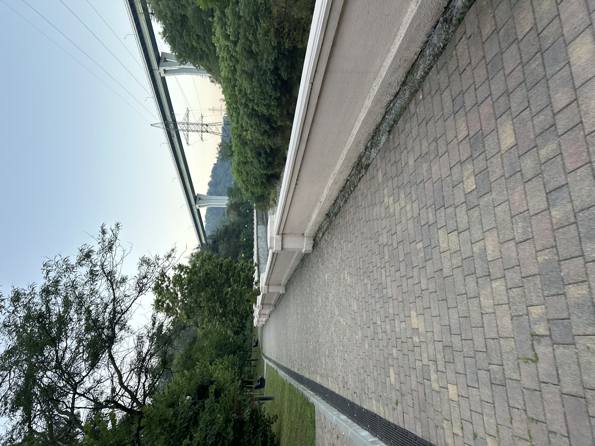 A paved walkway lined with a beige railing runs through a park-like area with mature trees. In the background, a large elevated highway bridge crosses over a gorge, with power lines and a transmission tower visible. The sky is clear and pale blue. The scene appears to be in Sochi, Russia, possibly near the Adler district, given the infrastructure and natural landscape.