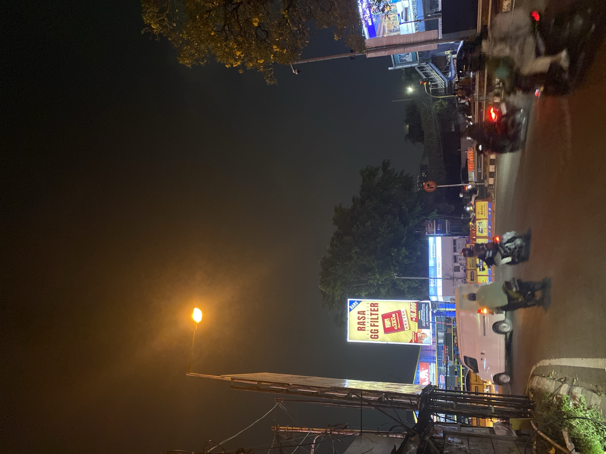 This image depicts a nighttime street scene in Tangerang, Indonesia, likely near a major intersection or thoroughfare. A bright streetlamp illuminates a hazy sky, casting a warm glow over the scene. In the foreground, a white van is driving on the road, with a motorcycle and its rider in front of it. Another motorcycle is visible to the right, also in motion. A large billboard advertising "GG Filter" cigarettes is prominently displayed on the left side of the image, featuring text in Indonesian and a graphic of a cigarette pack. Other illuminated billboards and signs for businesses like "Otrtv" and "Pos Pantau" are visible further down the street. Trees with yellow foliage are present on the right side of the image, and a pedestrian overpass with stairs and railings can be seen in the background. The overall impression is a bustling urban environment at night, with traffic and commercial activity.