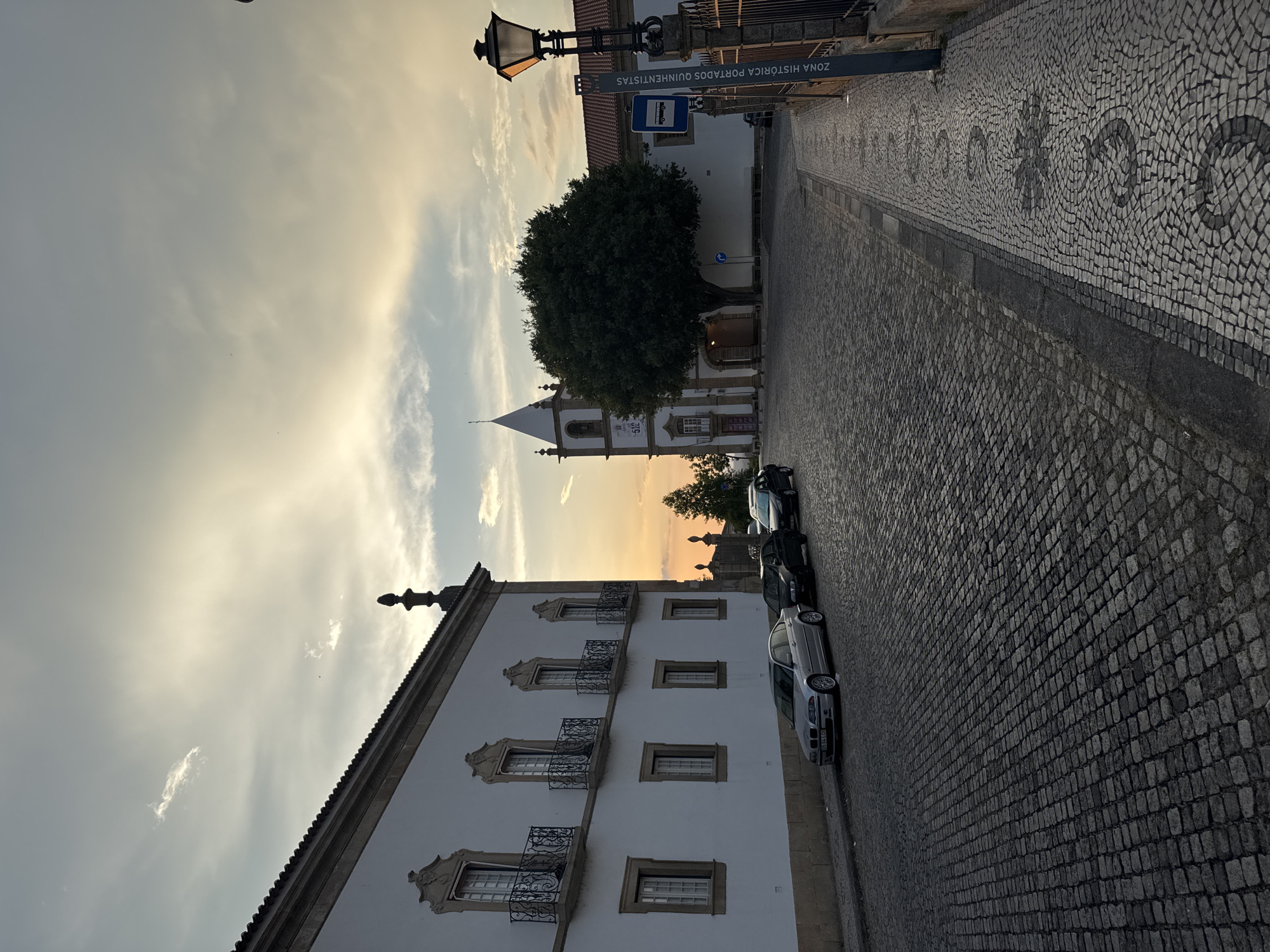 The image captures a view of a cobblestone street in Castelo Branco, Portugal, at dusk. The sky is filled with soft, diffused light from the setting sun, with scattered clouds. On the left, a white building with a stone base and ornate windows with iron balconies lines the street. Several cars are parked along the building's facade.
In the background, a church with a white steeple and a bell tower stands prominently. A large, leafy tree is situated in front of the church. To the right of the church, another building with a red-tiled roof is visible. A street lamp with a classic design illuminates the scene from the right side of the frame. A signpost with text, likely indicating a historical zone, is also present. The street itself is paved with cobblestones, with a decorative mosaic pattern on the sidewalk to the right. The overall atmosphere is serene and historic.