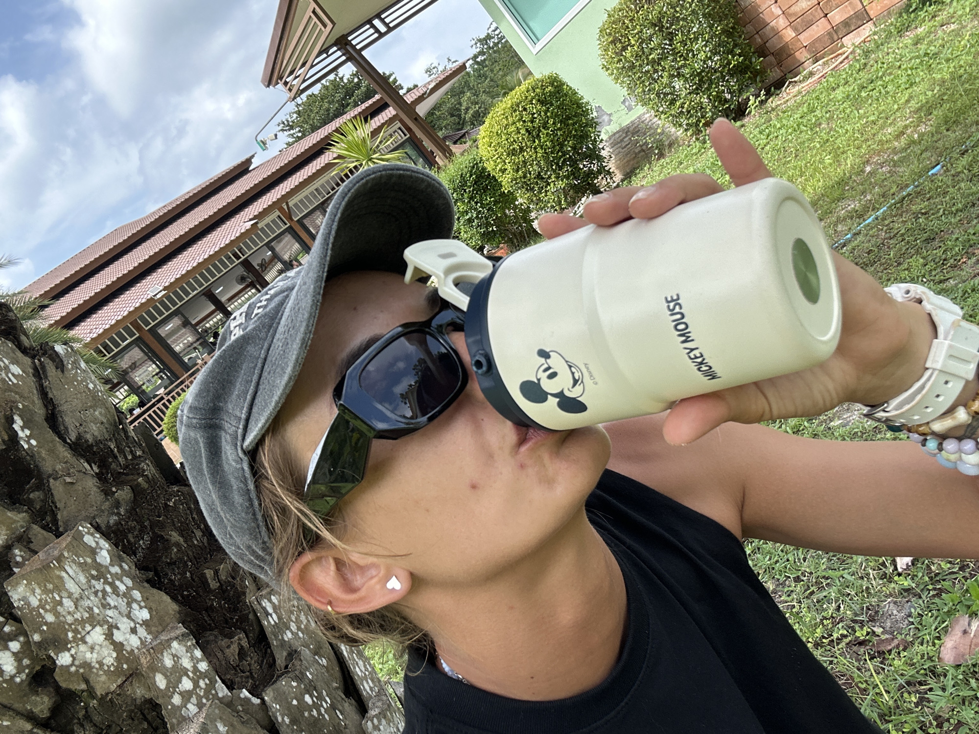 A woman wearing a grey baseball cap, dark sunglasses, and a black tank top is taking a selfie outdoors. She is holding a light-colored travel mug with Mickey Mouse branding up to her lips and appears to be drinking from it. Her left ear is visible, adorned with a small heart-shaped earring. A bracelet is visible on her right wrist. The background includes a large rock formation on the left, a building with a red tiled roof and a balcony on the right, and lush green trees and bushes. The setting is consistent with the outdoor environment of Ao Nang, Thailand, potentially near a resort or residential area, with a blue sky and scattered clouds visible overhead.