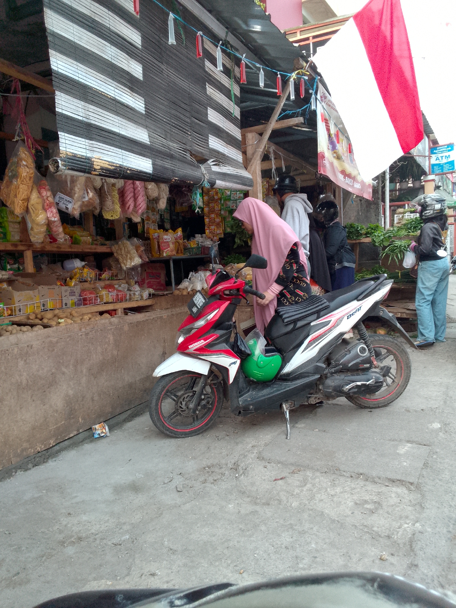 In Padalarang, Indonesia, a street-level scene captures a vendor stall with various goods displayed on shelves, including bags of snacks and what appear to be potatoes in the foreground. A red and white Honda Beat scooter is parked in front of the stall, with a green helmet placed on its seat. A woman wearing a pink hijab and a patterned long-sleeved shirt is interacting with the stall, possibly making a purchase. Behind her, a man in a grey hoodie and two other individuals are also present, one wearing a black helmet. Further in the background, a large Indonesian flag is visible, along with a sign for an ATM. The environment suggests a typical Indonesian marketplace or roadside stall.