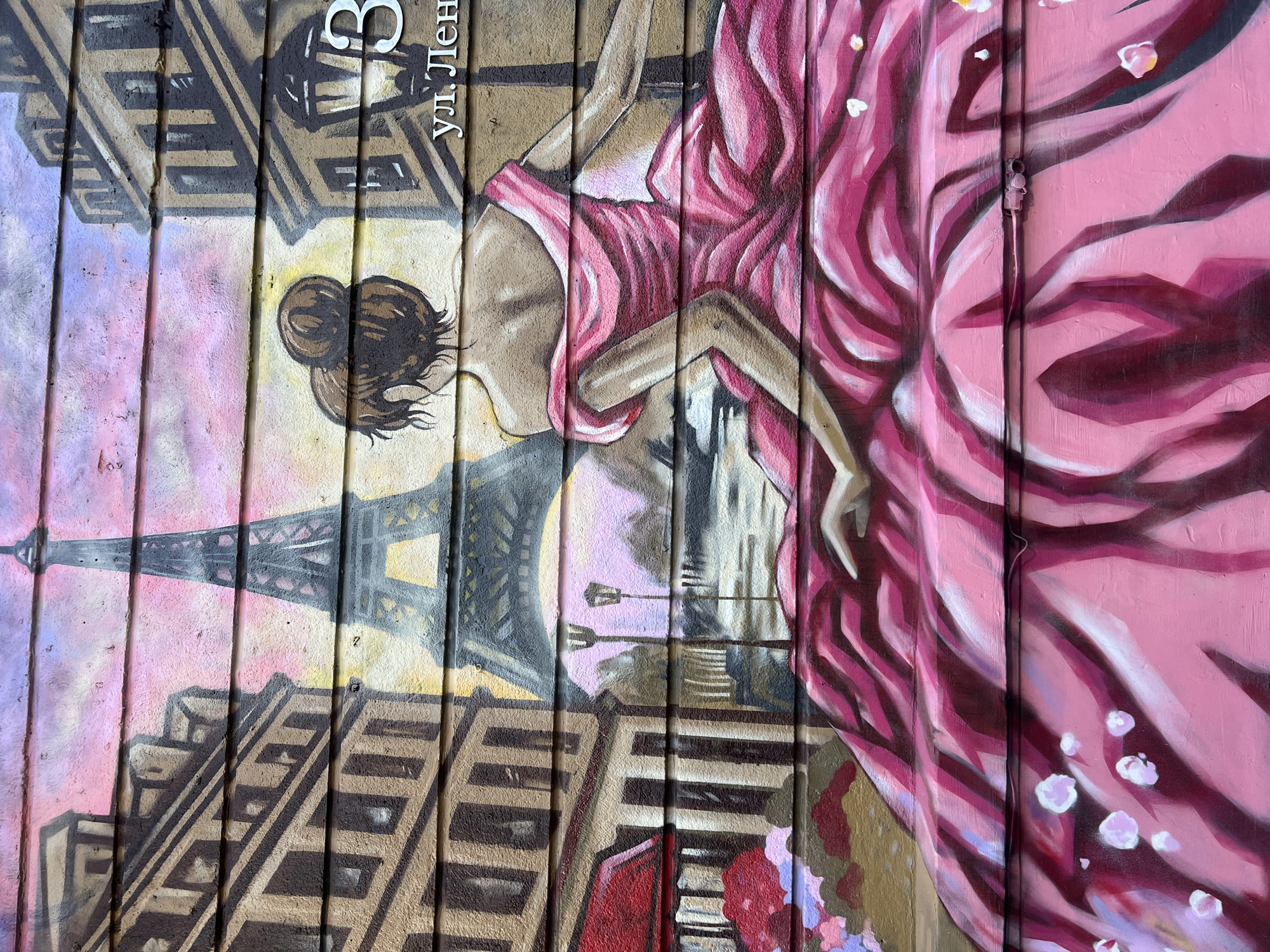 The image is a mural painted on a corrugated metal surface. The mural depicts a woman in a pink dress, viewed from the back, with her hair in a bun. The background includes the Eiffel Tower and buildings, suggesting a Parisian scene. The text "ул. Лен" and the number "3" are visible, indicating the location is 3 Lenina Street in Novosibirsk, Russia. The mural is painted in a loose, artistic style with visible brushstrokes.