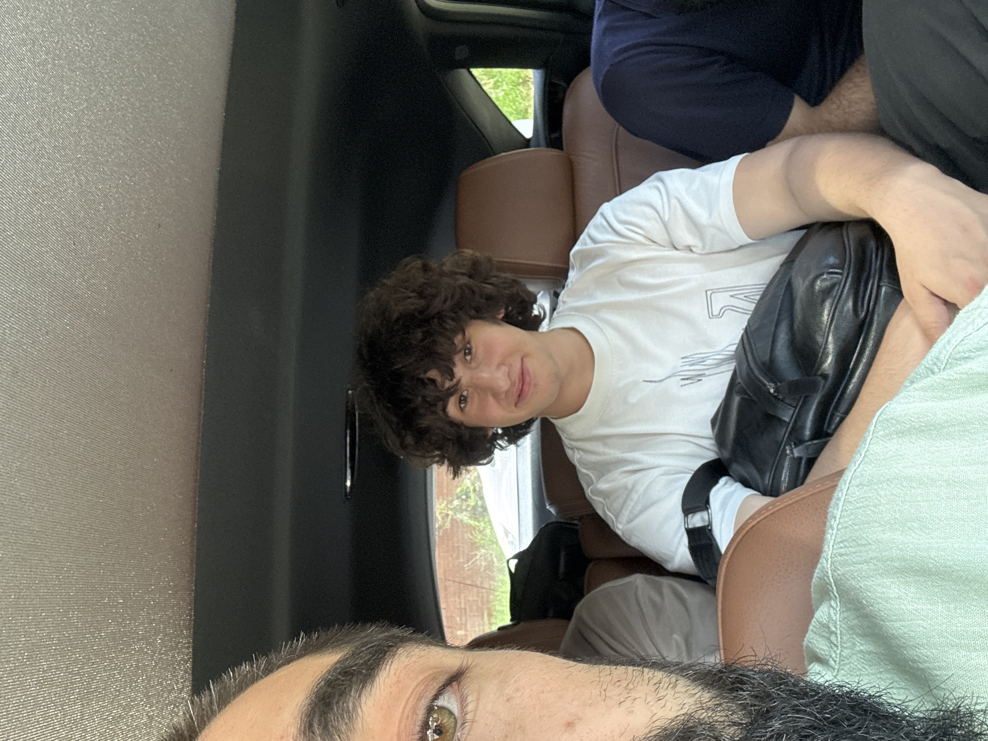 The image captures a group of individuals inside a vehicle, seemingly in motion. The foreground is dominated by the left side of a person's face, including a visible eye with an olive-green iris and a portion of a dark beard. This person is wearing a light green collared shirt.
In the mid-ground, a young male with curly brown hair is seated and looking towards the camera with a slight smile. He is wearing a white t-shirt with a graphic print. He is holding a black bag on his lap. Another person, wearing a dark navy shirt, is partially visible on the right side of the frame. The vehicle's interior features brown leather seats and a dark ceiling.
Through the windows, a blurred outdoor scene is visible, suggesting a daytime setting with foliage and a brick wall, hinting at a suburban or semi-rural environment. While the exact location within Novaya Adygeya, Russia, cannot be precisely determined from the visual cues, the overall context suggests a casual road trip or outing among friends or family.