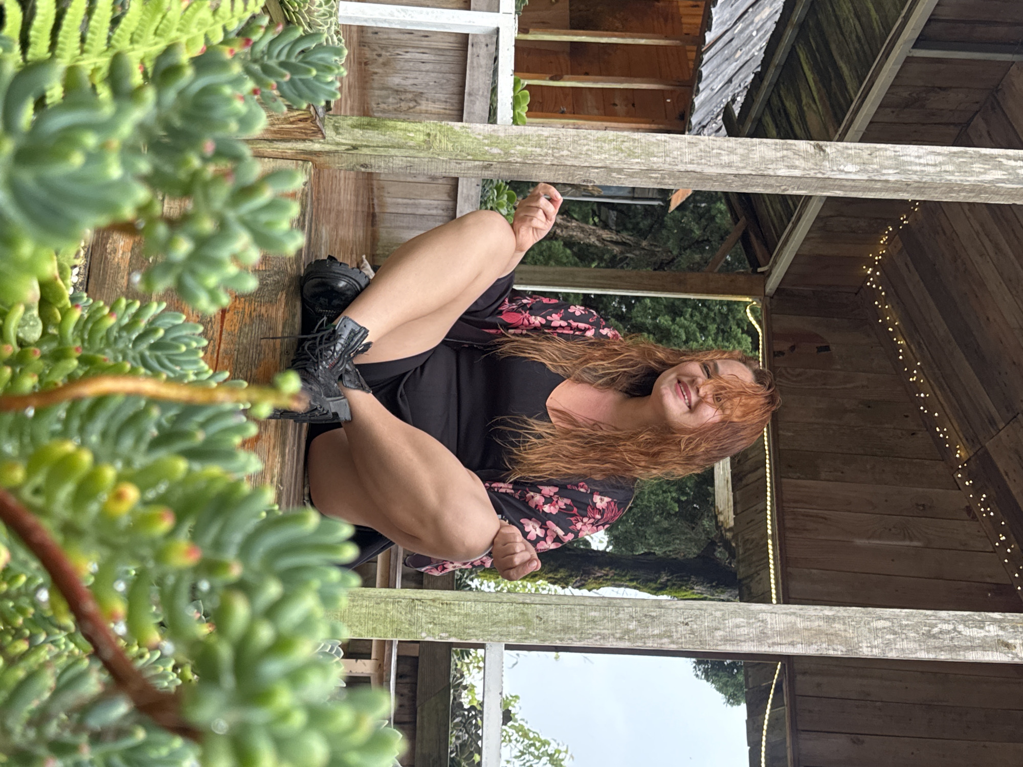 A Caucasian woman with long, auburn hair sits cross-legged on a wooden platform under a covered, rustic structure with exposed wooden beams and roof planks. She is wearing a black dress and a floral kimono with pink and red flowers. Her hands are in a meditative pose, with fingers lightly touching. In the foreground, a cluster of green succulents with red tips are in focus, obscuring the lower half of the woman's body. In the background, there is a wooden building with a corrugated metal roof, trees, and an overcast sky visible through the open sides of the structure. The overall environment appears to be outdoors, possibly a garden or nursery, in Lộc Quý, Vietnam, indicated by the presence of unique succulents and the rustic, tropical-style architecture.