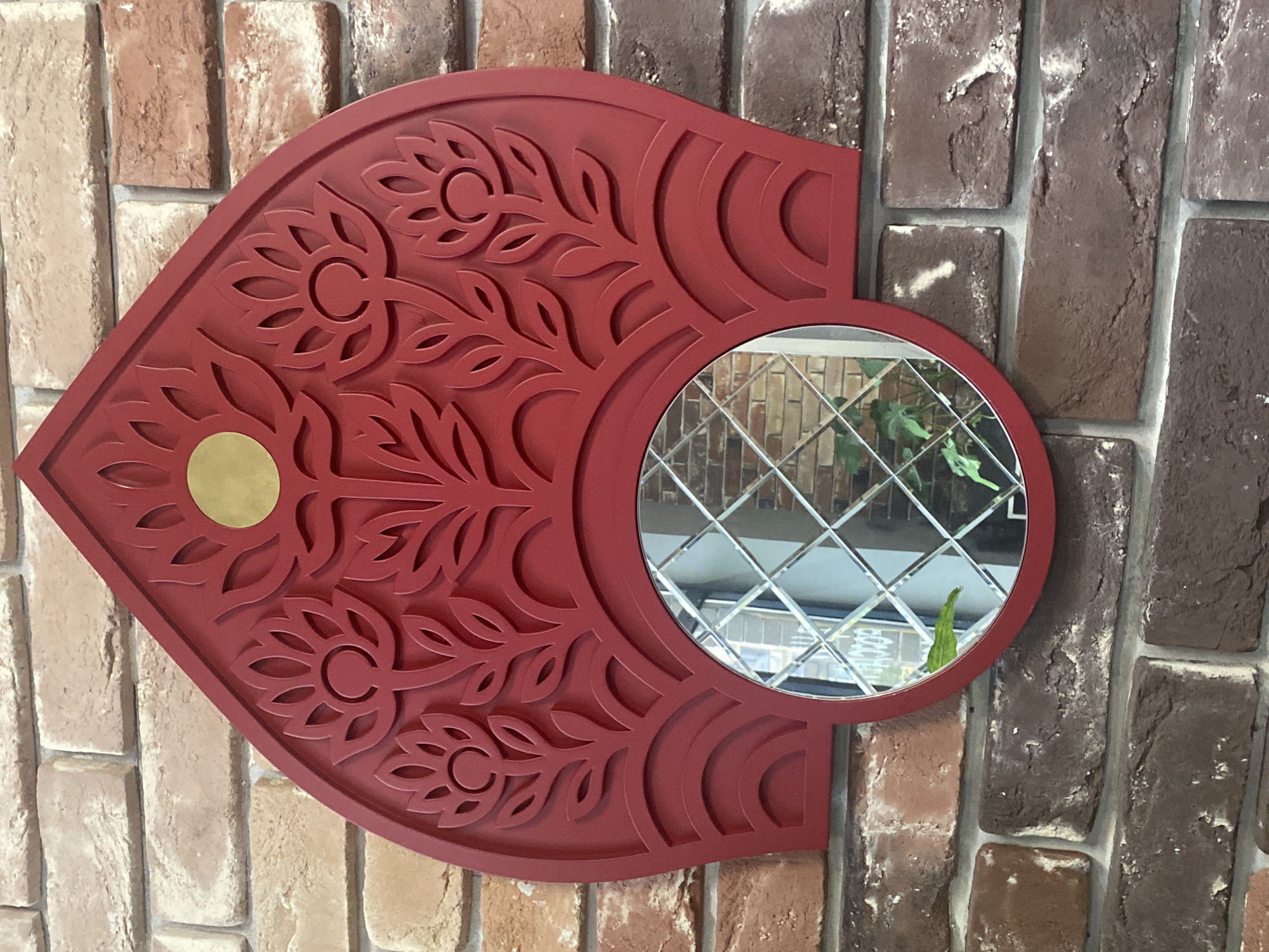 A red decorative mirror with a floral cutout design and a central circular mirror is mounted on a brick wall. The mirror is shaped like a kokoshnik, a traditional Russian headdress, with stylized flower and leaf motifs carved into the red frame. A small gold disc is positioned at the top center of the frame. The mirror's surface is beveled, creating a diamond-like pattern within the reflection. The brick wall exhibits variations in color, ranging from light tan to dark brown, with visible mortar lines. Plants are reflected in the mirror, suggesting an indoor environment. The mirror is located in the city of Mirny, Russia.