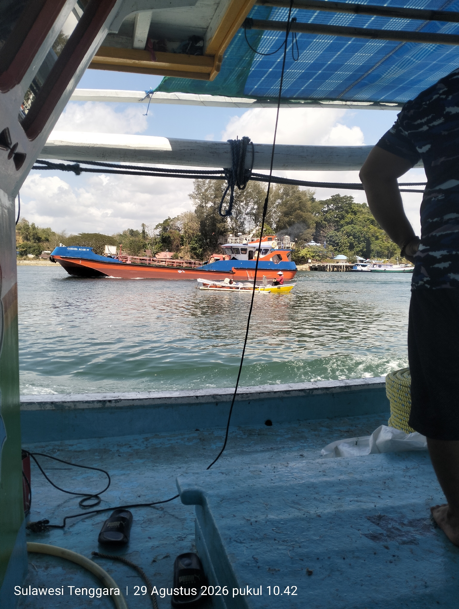The image depicts a view from the deck of a boat looking out at a body of water. In the distance, a large orange and blue barge with the name "CAHAYA ALAM IV" is visible. To the left of the barge, a smaller yellow boat with a person wearing a conical hat is also present. The water is choppy, indicating movement. The foreground shows the blue deck of the boat from which the photo is taken, with some black sandals and a yellow rope visible. In the upper right portion of the image, the arm and shoulder of a person wearing a dark blue shirt with a camouflage pattern are visible. Trees and some structures are visible on the shoreline in the background. The timestamp at the bottom indicates the location is Sulawesi Tenggara, Indonesia, on August 29, 2026, at 10:42 AM.