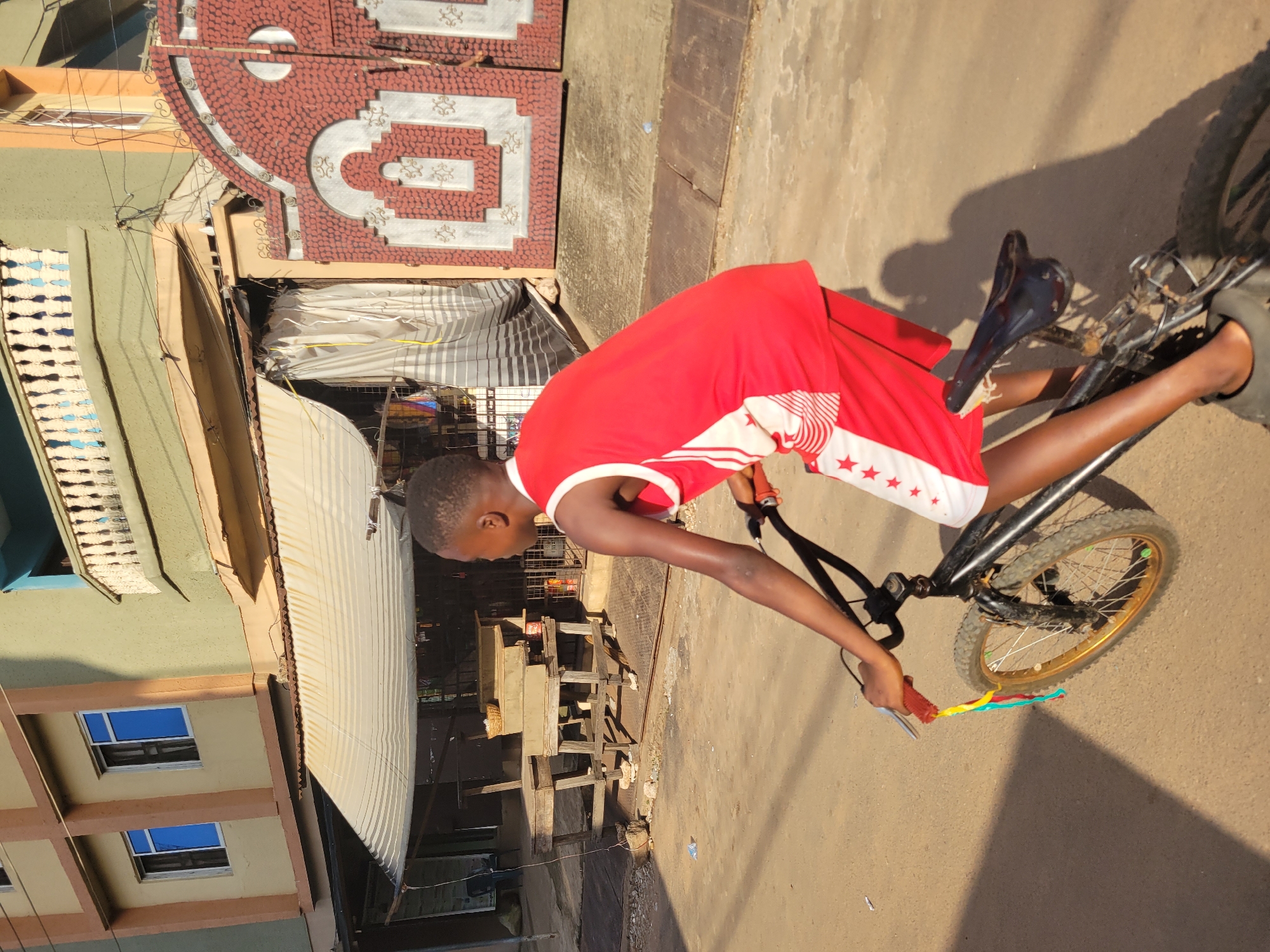 A young Black boy rides a bicycle in what appears to be a street in Onitsha, Nigeria. The boy wears a red and white tank top with red shorts that have white star accents. He is facing to the right of the frame, looking away from the camera. The bicycle is black with gold rims on the wheels and has colorful streamers hanging from the handlebars. The boy's left foot is on the pedal, and he appears to be in motion.
The environment is urban, with buildings visible in the background. To the left, there is a multi-story building with beige and brown facades, featuring several windows with blue glass. To the right, a green building with a white balcony railing is visible, adjacent to a large, ornate red and silver gate. In the middle ground, there is a covered stall or shop with a striped awning and white curtains. Wooden tables and benches are set up in front of this stall, and a basket is placed on one of the tables. The ground is a dusty, sandy surface, suggesting a typical street environment. Sunlight casts shadows across the scene, indicating it is likely late afternoon or early morning.
