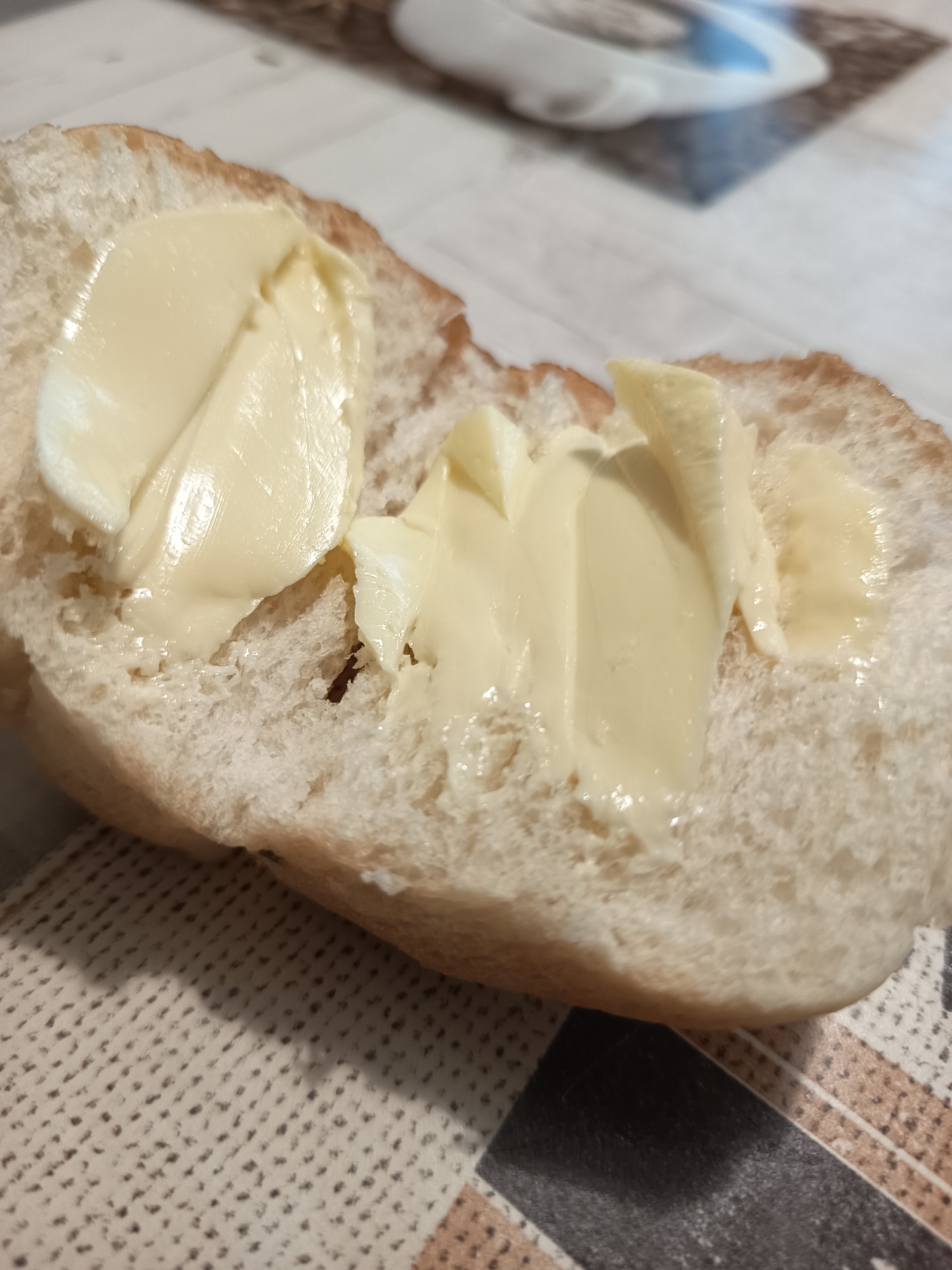 A close-up, eye-level shot shows a halved bread roll generously spread with butter, resting on a patterned placemat. The butter is a light yellow and has been applied in thick, uneven strokes, creating a creamy texture. The bread is soft and white on the inside, with a slightly browned crust. In the blurred background, a table surface is visible, along with a portion of a white object with blue accents. The lighting is overhead, casting soft shadows. The image does not contain any specific information about the city of Izmail, Ukraine.