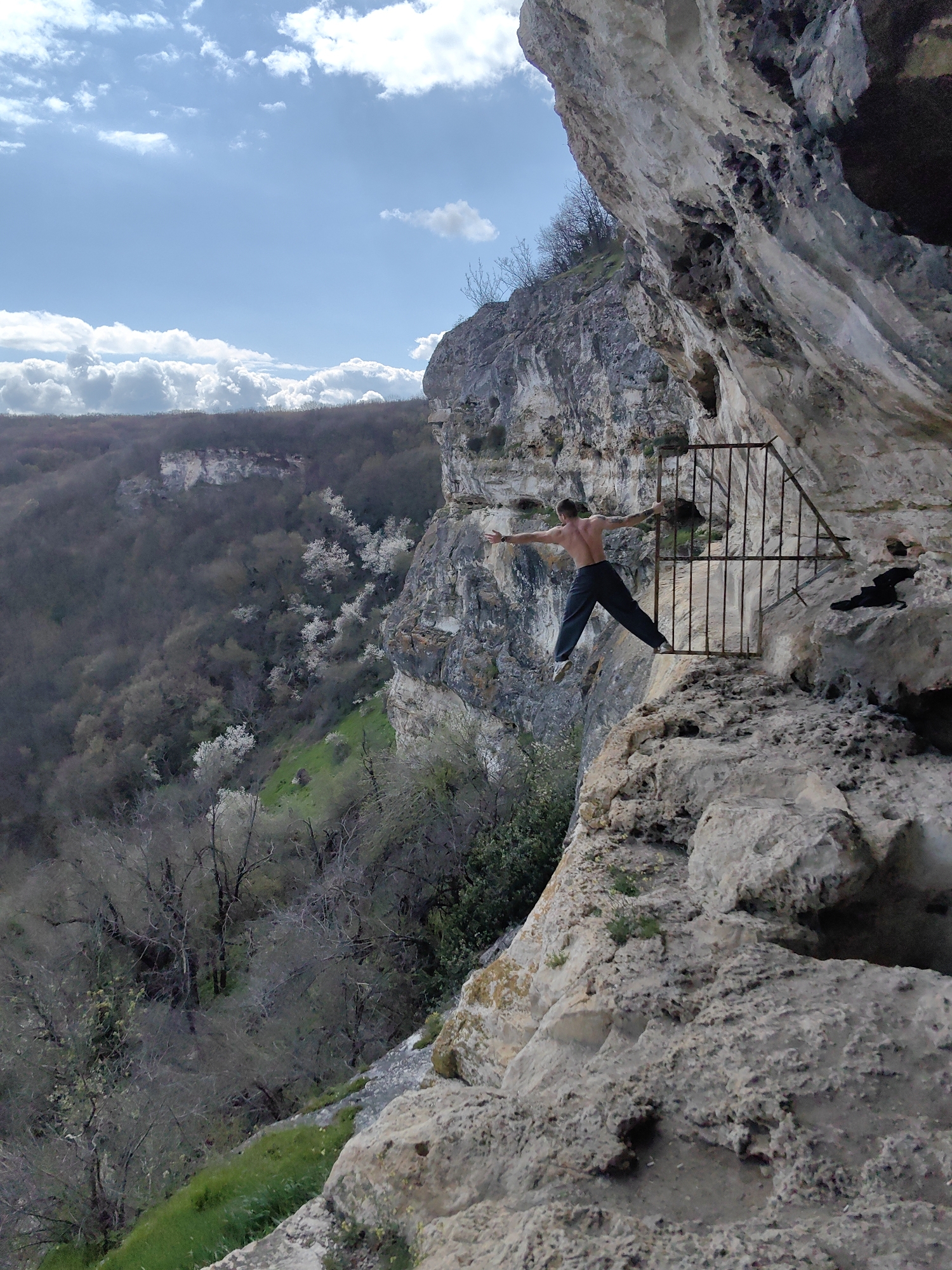 A man with a bare torso stands precariously on the edge of a rocky cliff, grasping onto a rusty, metal gate. The location is in the city of Shumen, Bulgaria. He is wearing dark pants and light-colored shoes. The gate appears to be a barrier or enclosure attached to the side of the cliff face. Behind him, the landscape features a forested valley with trees interspersed with patches of white blossoms, potentially indicating the spring season. The sky is partly cloudy with a mix of blue and white. The immediate foreground consists of a textured, light-colored rock surface, while the background shows a panoramic view of the hilly terrain.