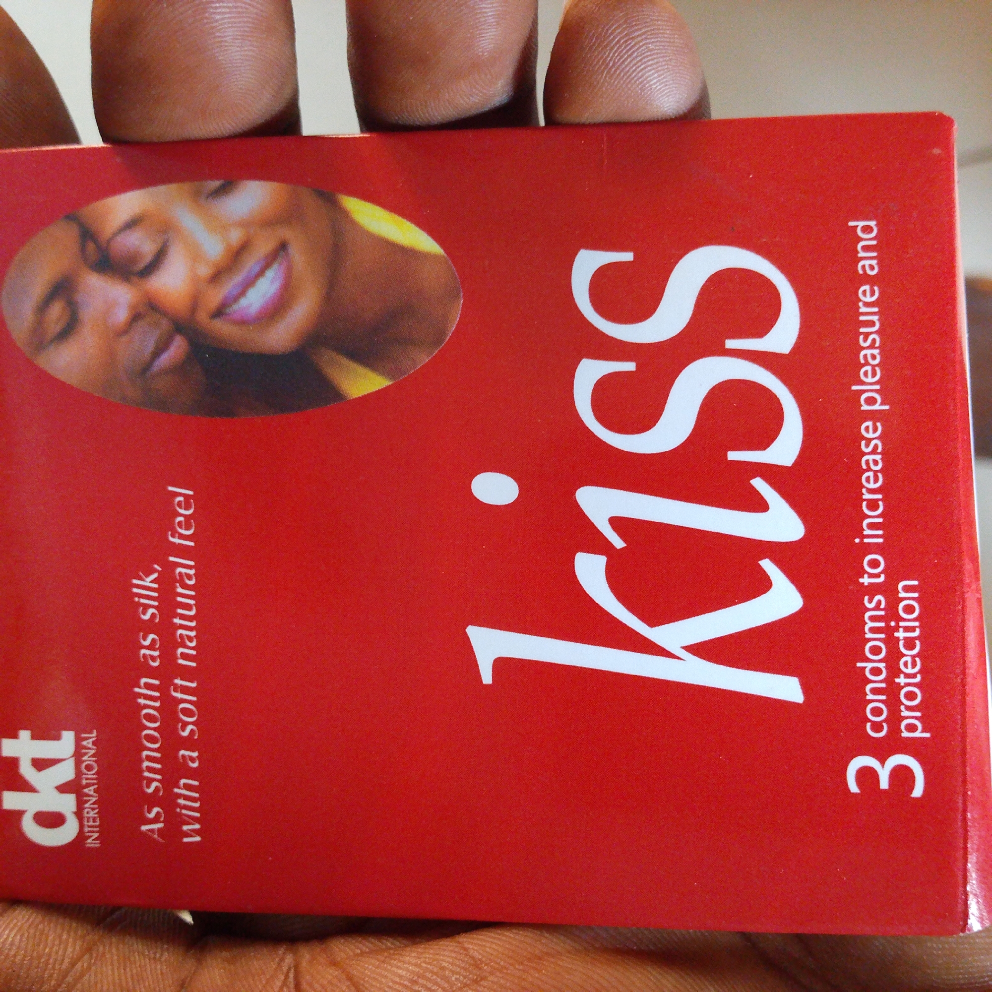 A close-up, eye-level shot captures a red box of "Kiss" condoms, held by fingers. The box features an oval image of a couple embracing, their faces close together. The brand name "Kiss" is prominently displayed in large white letters on the right side of the box. To the left of the brand name, smaller white text reads "3 condoms to increase pleasure and protection". Below the oval image, more white text states "As smooth as silk, with a soft natural feel". In the bottom left corner of the box, a white logo with the letters "dkt" and the word "INTERNATIONAL" beneath it is visible. The background is out of focus, suggesting an indoor environment with soft lighting. The image is sharp and clear, showcasing the product packaging in detail.