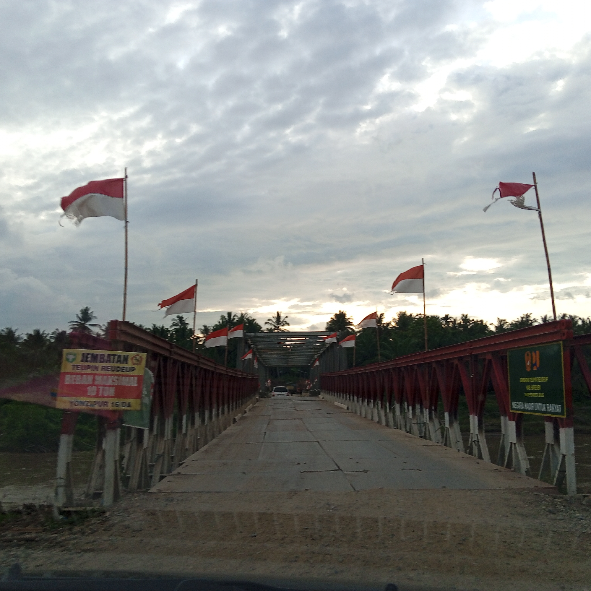 The image depicts a straight-on, low-angle view of the Teupin Reudeup bridge in Awegeutahpaya, Indonesia, from the perspective of a vehicle entering or traversing it. The bridge features a red-painted metal truss structure on both sides, supporting a roadway constructed from grey metal plates or concrete slabs, some showing signs of patching and wear. Numerous red and white flags, characteristic of the Indonesian national flag, are affixed to poles along both sides of the bridge, many appearing wind-blown and some visibly tattered.
On the left side of the bridge, a yellow and red sign with black text indicates "JEMBATAN TEUPIN REUDEUP," states a "BERAN MAKSIMAL 10 TON" (maximum load 10 tons), and mentions "YONZIPUR 16 DA" (Combat Engineer Battalion 16th Detachment/Area). Beneath the bridge on the left, a brown river is visible, flanked by dense green vegetation.
On the right side of the bridge, a green and yellow sign with white text also identifies the "JEMBATAN TEUPIN REUDEUP," mentions "ACEH BEREGEN," and specifies "10 DESEMBER 2016," followed by the slogan "NEGARA HADIR UNTUK RAKYAT" (The State is Present for the People).
In the distance, a white car is visible on the bridge, approaching the viewer. Beyond the bridge's structure, the surrounding environment consists of dense tropical foliage, primarily palm trees, extending under an overcast sky with scattered, bright clouds.