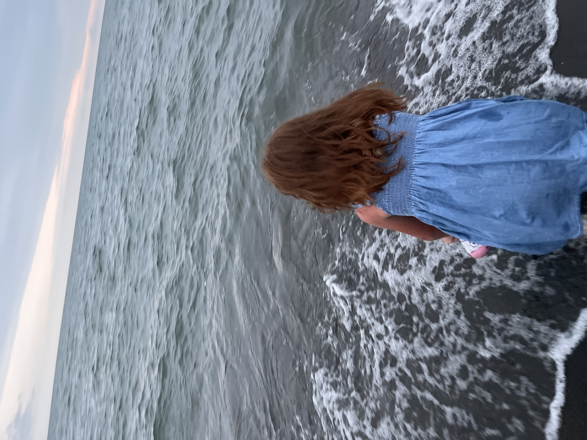 A young girl with long, reddish-brown hair is crouched at the edge of the sea, her back to the camera. She is wearing a blue denim dress with a smocked bodice and white sneakers with pink accents. The water is a deep grey-blue, with gentle waves lapping at the dark, sandy shore. Foam and ripples from the waves are visible around her. In the background, the sky is a soft, pale blue with streaks of pink, suggesting it is either dawn or dusk. The overall scene conveys a sense of peaceful engagement with nature.